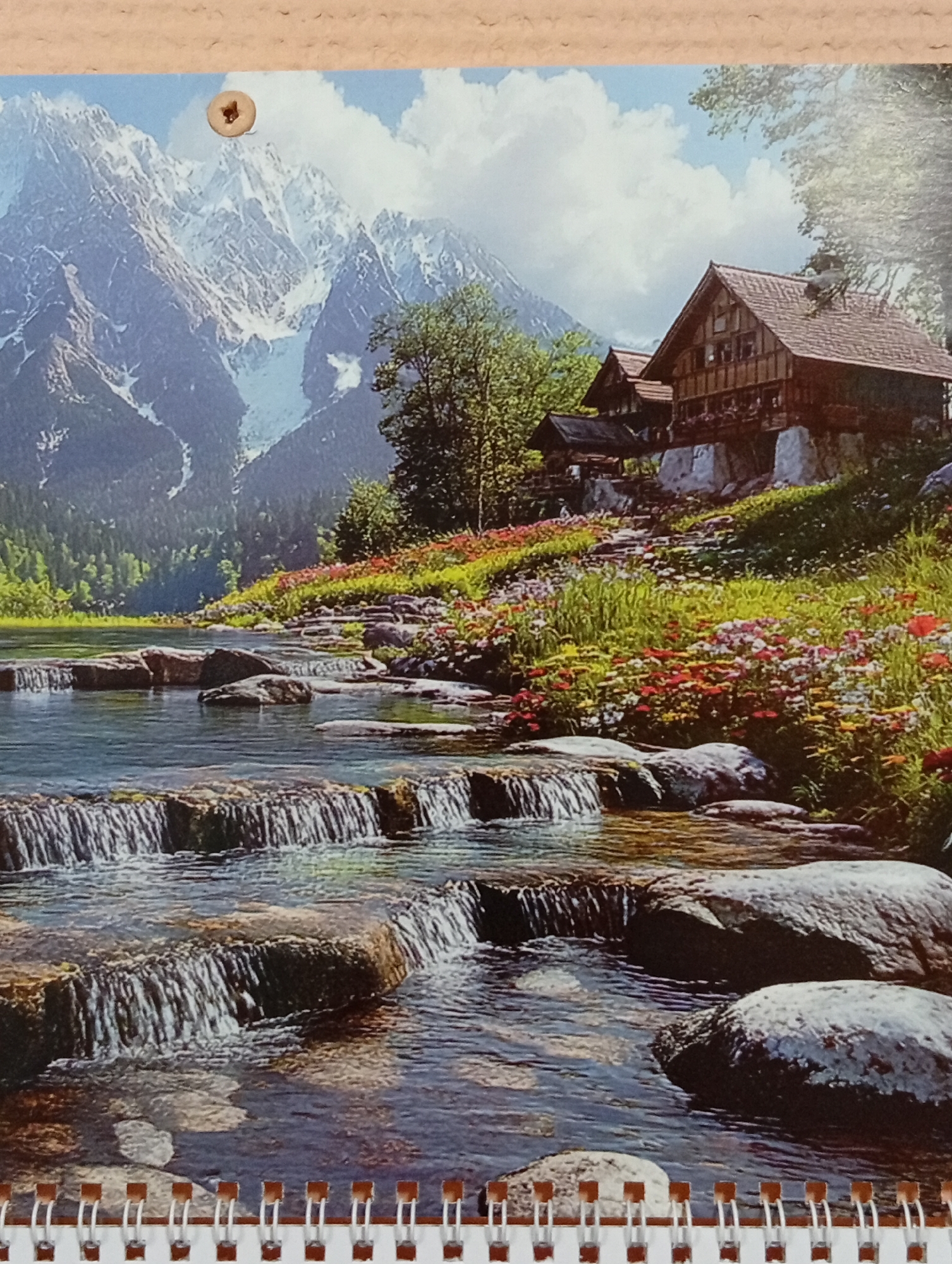 The image depicts a scenic mountain landscape with a charming wooden cabin nestled on a hillside. A clear, rocky stream with small waterfalls flows through the foreground, surrounded by lush green grass and a vibrant array of colorful wildflowers. Majestic snow-capped mountains rise in the background, partially obscured by fluffy white clouds against a bright blue sky. The cabin itself is a multi-story structure with a prominent pitched roof, featuring balconies and windows, suggesting it is a rustic dwelling. The overall scene conveys a sense of tranquility and natural beauty. The image is a printed depiction of this scene, with a small circular fastener visible at the top center of the print, securing it against a textured, light-colored backing material. This fastener suggests the print is part of a larger item, such as a calendar or a binder. The bottom edge of the image shows a spiraling wire binding, typical of a notebook or calendar, with small, rectangular protrusions. The image does not contain any discernible elements that would place it specifically in Pushkino, Russia.