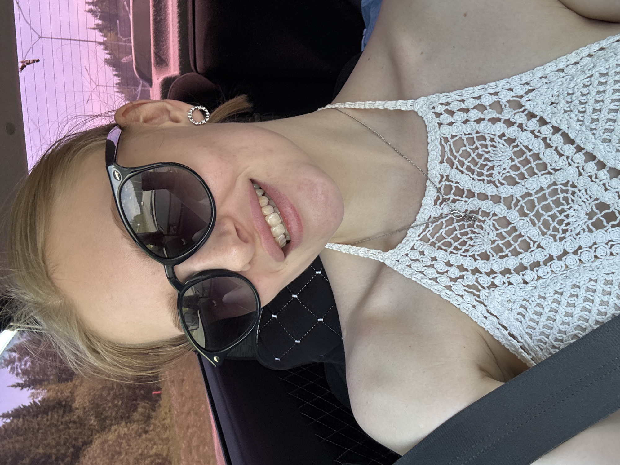 A selfie of a woman with fair skin, wearing black sunglasses and a white crocheted top, seated in a vehicle with a seatbelt fastened. The earring in her left ear is a circular design. A silver necklace is visible on her chest. The background shows a view of trees and foliage seen through the vehicle window, suggesting an outdoor or roadside location. The location is specified as Perm, Russia. The interior of the vehicle includes a black seat with diamond stitching.
