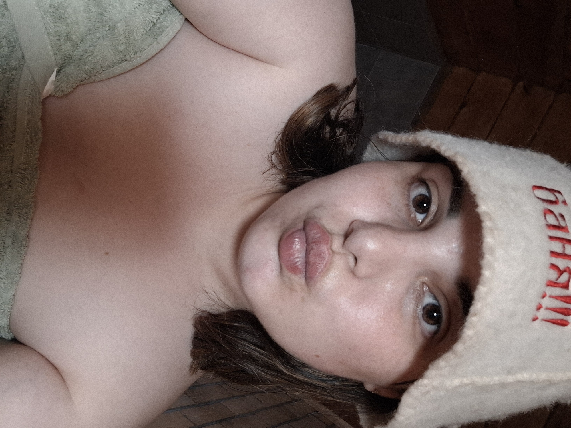 A person with light skin, brown hair, and a sauna hat is posing in a sauna in Амзя, Russia. The hat has red Cyrillic text on it. The person is partially covered by a green towel. The background consists of wooden planks and gray tiles. The person's lips are pursed.