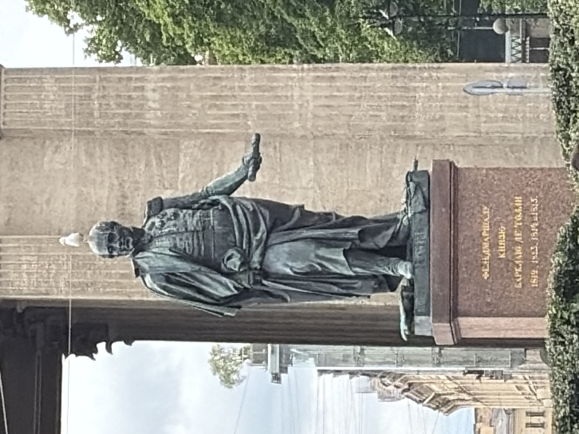 A full shot captures a bronze statue of a man in military attire, with a seagull perched on his head. The statue stands on a multi-tiered granite pedestal inscribed with gold lettering in Cyrillic. The man is depicted in a flowing robe over a military uniform, with one arm extended outward. Behind the statue, a colossal fluted column rises, part of a larger architectural structure. To the left of the statue, a building with multiple windows and ornate details is visible. In the background, lush green trees and a cloudy sky frame the scene. The statue is located in St. Isaac's Square in Saint Petersburg, Russia, and it depicts Field Marshal Mikhail Kutuzov.