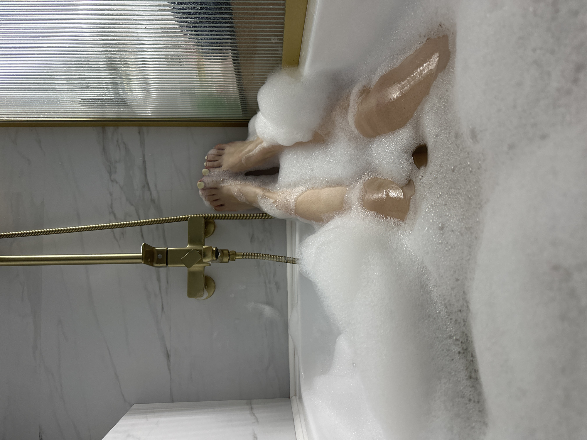 A photograph depicts a bathtub filled with white bubbles, partially obscuring two individuals. Two sets of feet with painted yellow toenails are visible protruding from the foam. A gold-colored shower fixture is attached to a white marble-tiled wall. A glass shower door with horizontal ridges is visible in the background. The image suggests an indoor bathroom setting but does not establish a location within the city of Chaikovsky, Russia.