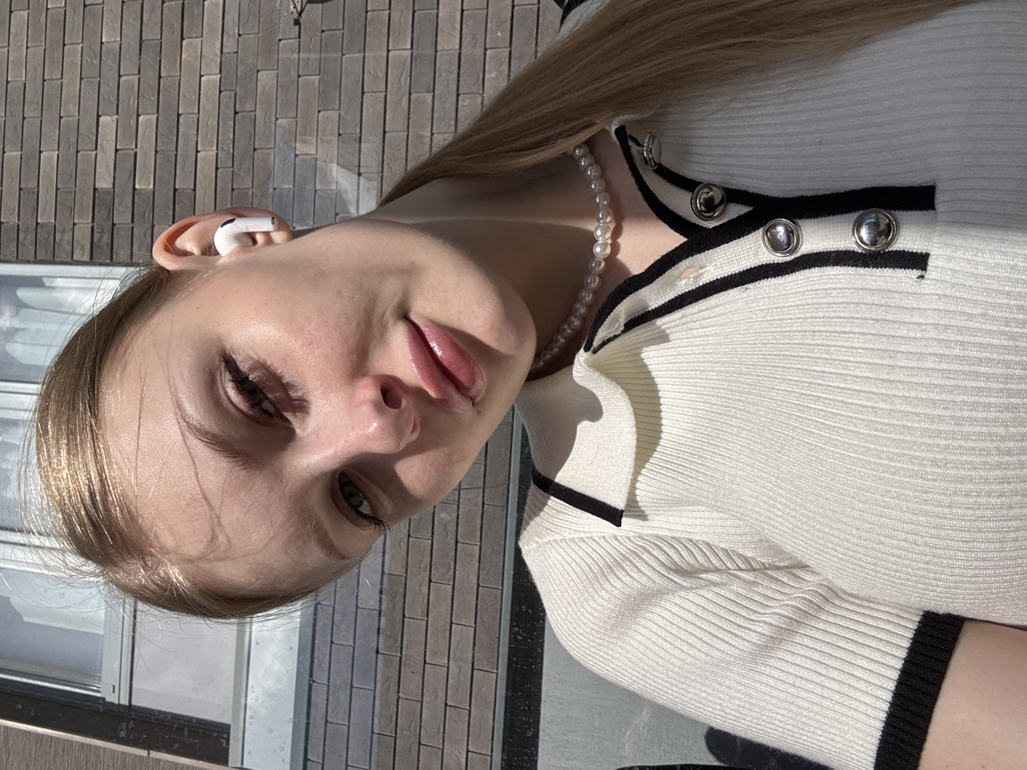 A young woman with blonde hair pulled back in a ponytail and wearing a white ribbed polo shirt with black trim and a pearl necklace poses for a selfie. She has a white earbud in her right ear and her left eye is looking directly at the camera. Her lips are slightly parted. The background consists of a brick wall and a window. This image was taken outdoors. Based on the provided image and context, it is impossible to determine the location within the city of Perm, Russia.