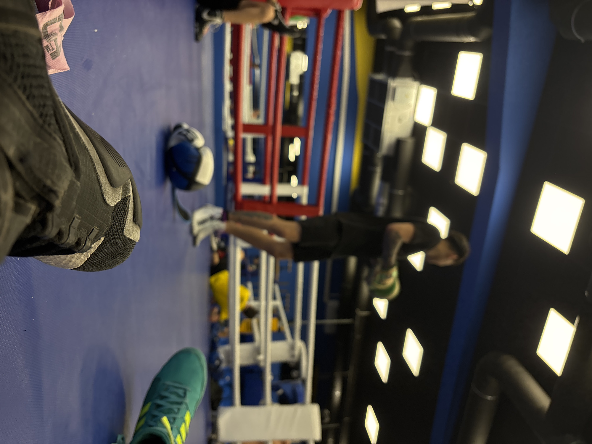 This image captures an indoor gym scene in Sofia, Bulgaria, seen from a slightly upward angle, with a focus on the floor and activities within a boxing ring. The perspective is from a person wearing athletic shoes.
In the foreground, partially cropped on the left, is a dark grey or black athletic shoe with a textured sole. To its right, another foot is visible, wearing a teal-colored athletic shoe with prominent yellow stripes and laces. A crumpled pink fabric item, possibly a towel, with faint white text (appearing as "TM" or a logo fragment) rests on the blue mat between the shoes.
The blue mat extends into the midground, leading to a boxing ring with a red frame, white ropes, and blue matting inside. A blue and white medicine ball lies on the mat near the boxing ring's edge. Inside the ring, a person in a black t-shirt and dark shorts is seen in a plank-like position, holding a small, light-green medicine ball with both hands. Their face is blurred or obscured, and they appear to be mid-exercise. Another person's legs and feet are visible in the far background, partially obscured by the ring structure.
The background features dark walls or ceiling with multiple rectangular, bright white light fixtures, illuminating the gym space. The overall setting is clearly an active training environment, likely during a workout session.