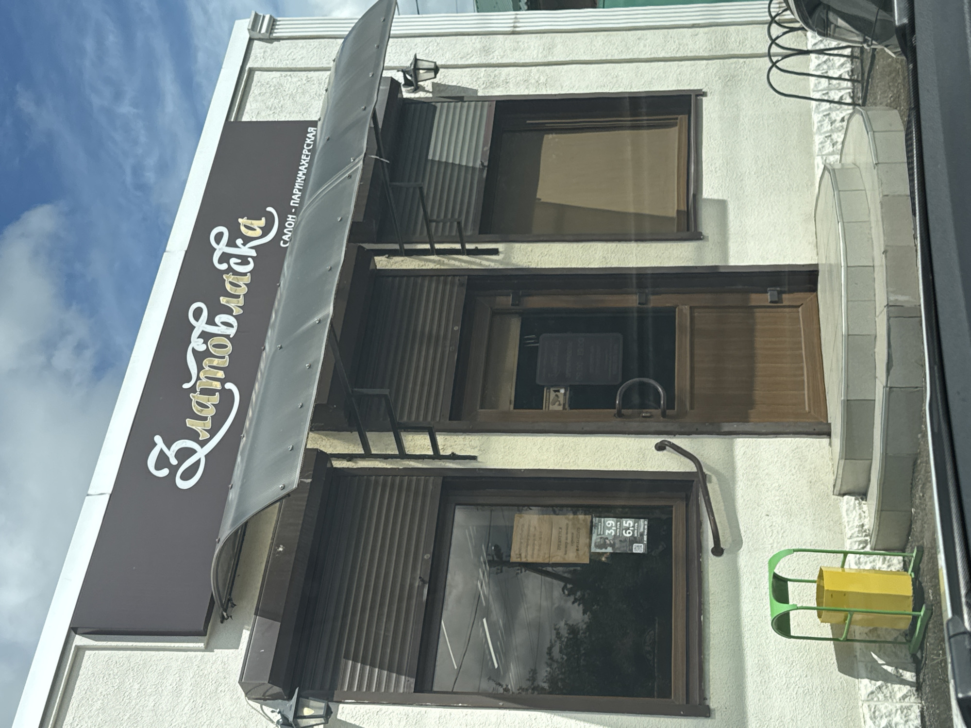 The image shows a three-story building in Yelizavetinskaya, Russia. The sign on the awning reads "Златовласка", with "САЛОН - ПАРИКМАХЕРСКАЯ" (salon - hairdresser) written below. The building has brown framed windows. The two lower windows have what appear to be security shutters partially closed. The second story window has a small sign inside. A metal handrail is located below the second-story window and a green and yellow trash bin is located to the right of the bottom window.