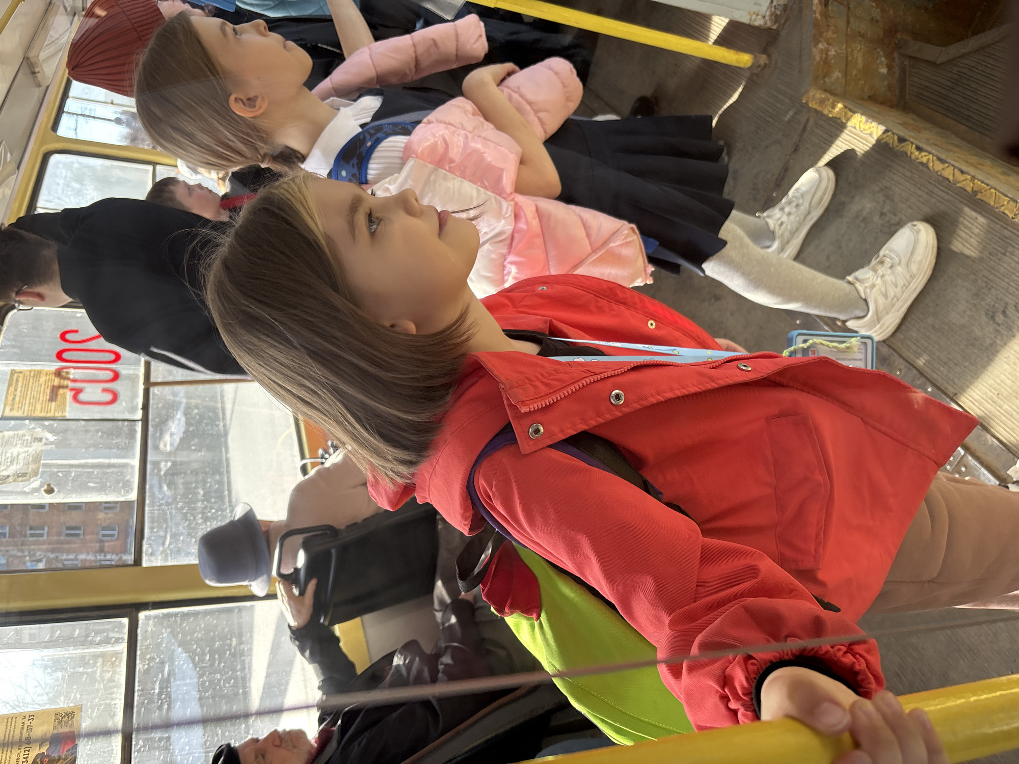 The image captures the interior of a public transport vehicle, likely a tram, in Izhevsk, Russia. Two young girls are present. One is wearing a red jacket and holding onto a yellow handrail. Another girl, wearing a dark dress with a white collar and a pink jacket, sits beside her. Other passengers are visible in the background, including one wearing a grey hat. The environment includes the tram's windows displaying urban scenes, advertisements, and other passengers. The tram’s floor shows signs of wear.