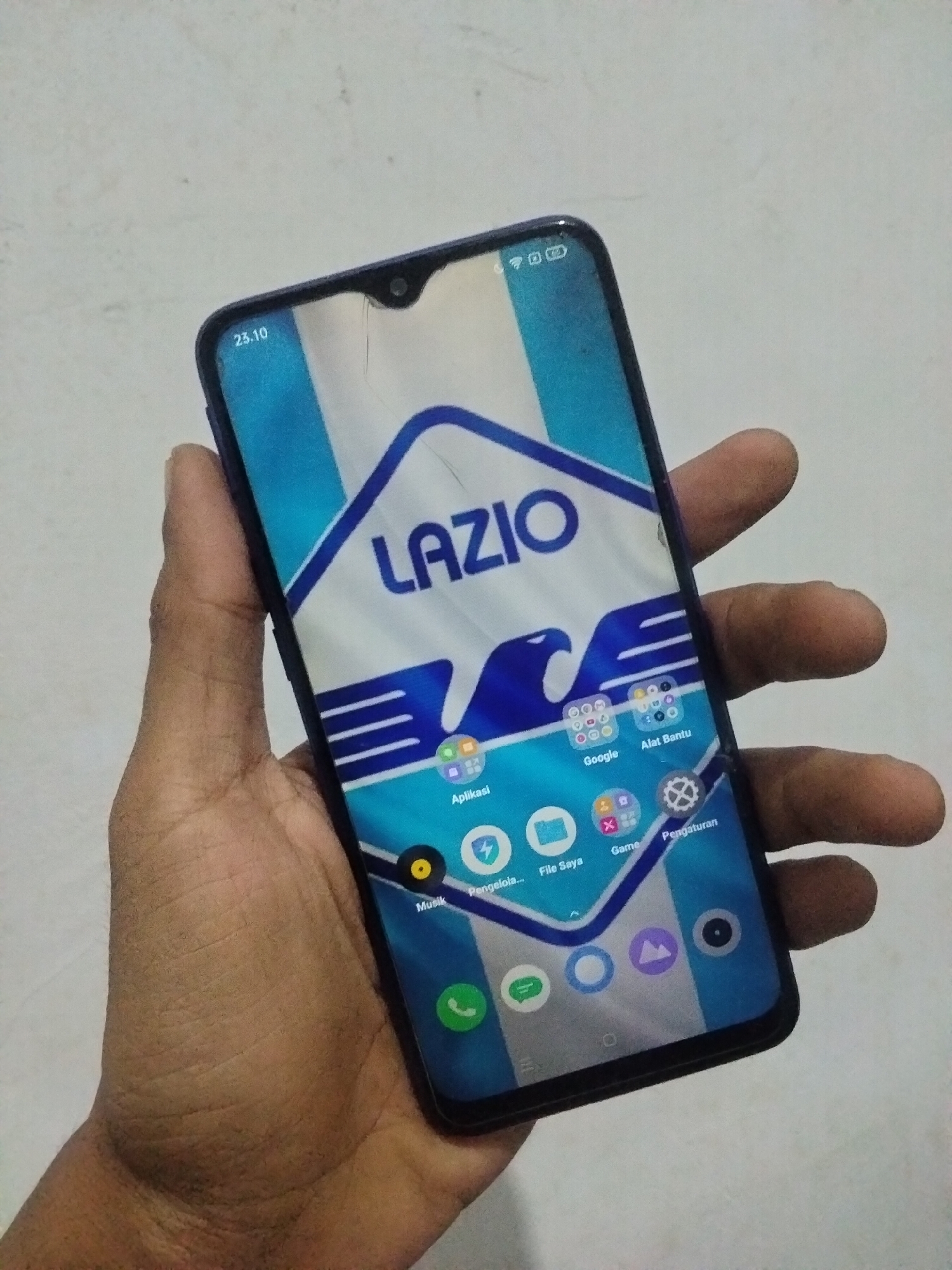 A person's hand is holding a smartphone with a blue and white wallpaper displaying the word "LAZIO" and an eagle graphic. The phone screen shows various app icons and status indicators. The background is a plain, light-colored surface. There are no visible indicators of the city of Antirogo, Indonesia, in this image.