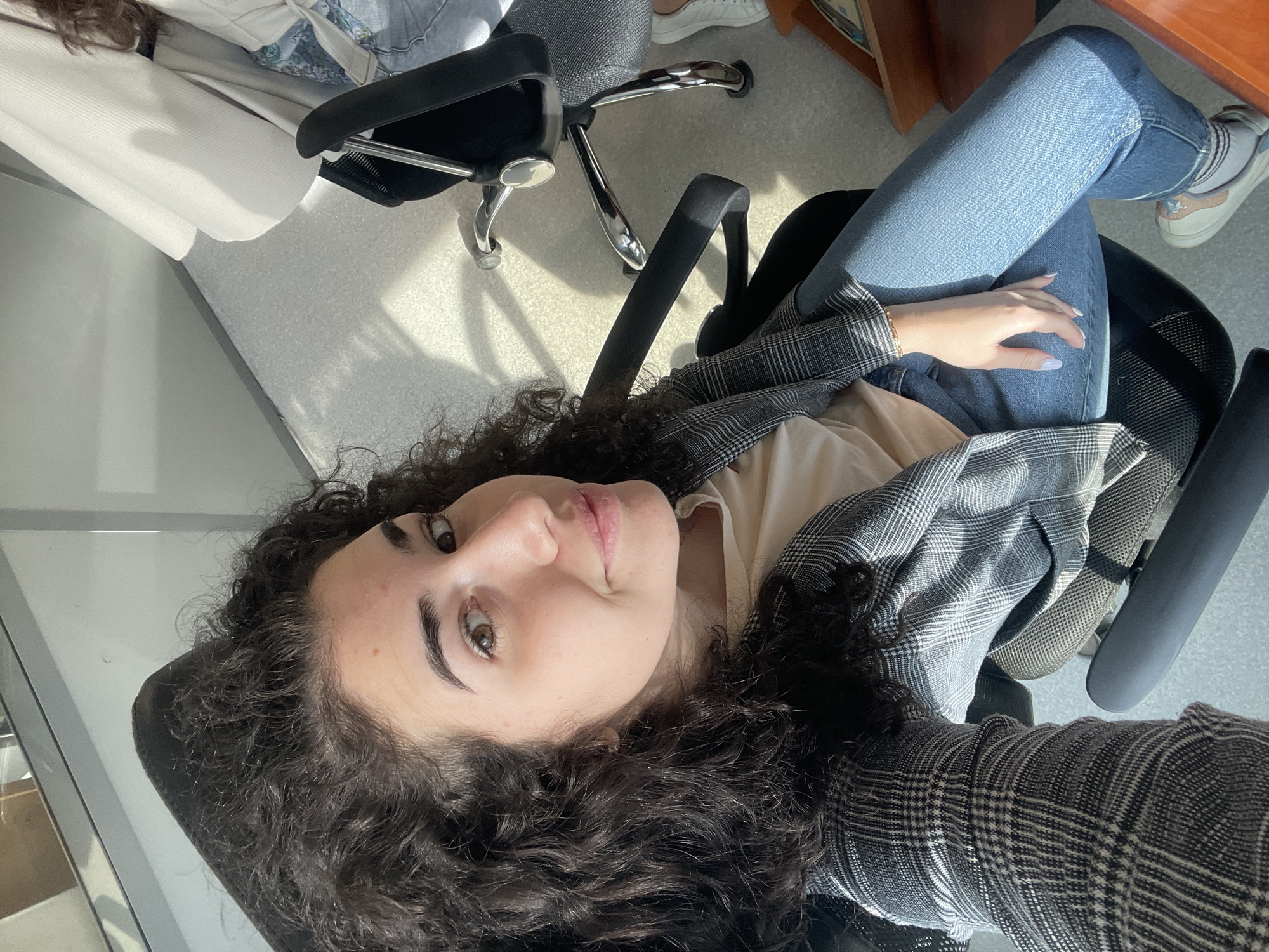 A selfie taken from above of a woman with curly dark hair, light skin, and brown eyes. She is sitting in a black office chair, wearing a light-colored shirt, blue jeans, a gray and white plaid blazer, and white sneakers. Her right hand rests on her leg. Another black office chair is partially visible to the left of her chair. The location appears to be indoors, with a light-colored floor and a window to the left. Sofia, Bulgaria cannot be determined from the information available.