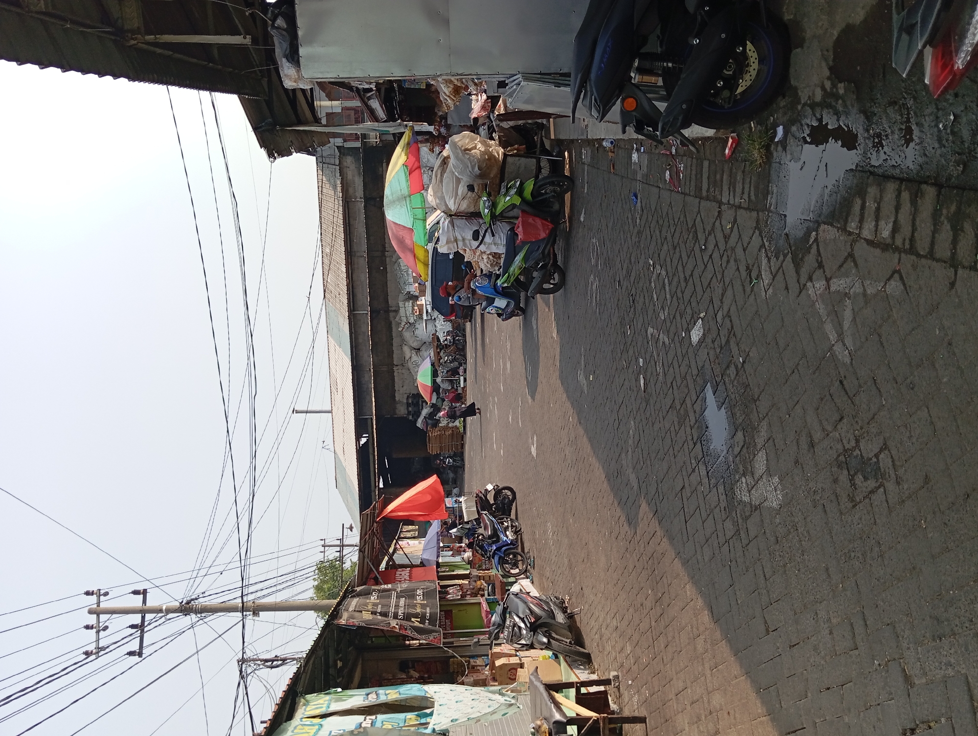 The image depicts an outdoor market scene in Kepulungan, Indonesia, with a focus on a paved walkway lined with stalls. The time of day appears to be late morning or early afternoon, given the angle of the sunlight casting shadows.
On the left side of the walkway, several stalls are visible, some with awnings and merchandise displayed. One stall has a table with boxes and what appears to be food items. A dark gray motorcycle is parked in front of this stall. Further down the walkway, another motorcycle, this one blue, is parked alongside a stall displaying signage. A bright red umbrella is unfurled above a stall behind the motorcycles.
The walkway itself is paved with dark interlocking bricks, with some areas appearing wet or having puddles. Scattered debris is visible on the ground, including paper and plastic.
In the mid-ground, a vibrant, multi-colored umbrella (green, yellow, red) is set up over a cart laden with large, white plastic-wrapped bundles. A person on a blue scooter is stopped near this cart, and another person is standing nearby. A green scooter is parked beside the cart, with a red bag hanging from its handlebars. A dark-colored minivan is parked behind these elements. Further into the background, more stalls and structures are visible, including what looks like a warehouse-style building with a corrugated metal roof. Several people are seen milling about in the distance, and more motorcycles are parked.
On the right side of the image, the edge of another stall structure is visible, with a large metal storage unit or refrigerator. A dark-colored motorcycle is parked in the foreground on the right, with its rear wheel and exhaust pipe clearly visible. More debris is scattered along the right edge of the walkway.
Above the market, a complex network of power lines and utility poles crisscrosses the sky, indicating the presence of electrical infrastructure in the area. The sky is clear and light, suggesting fair weather.