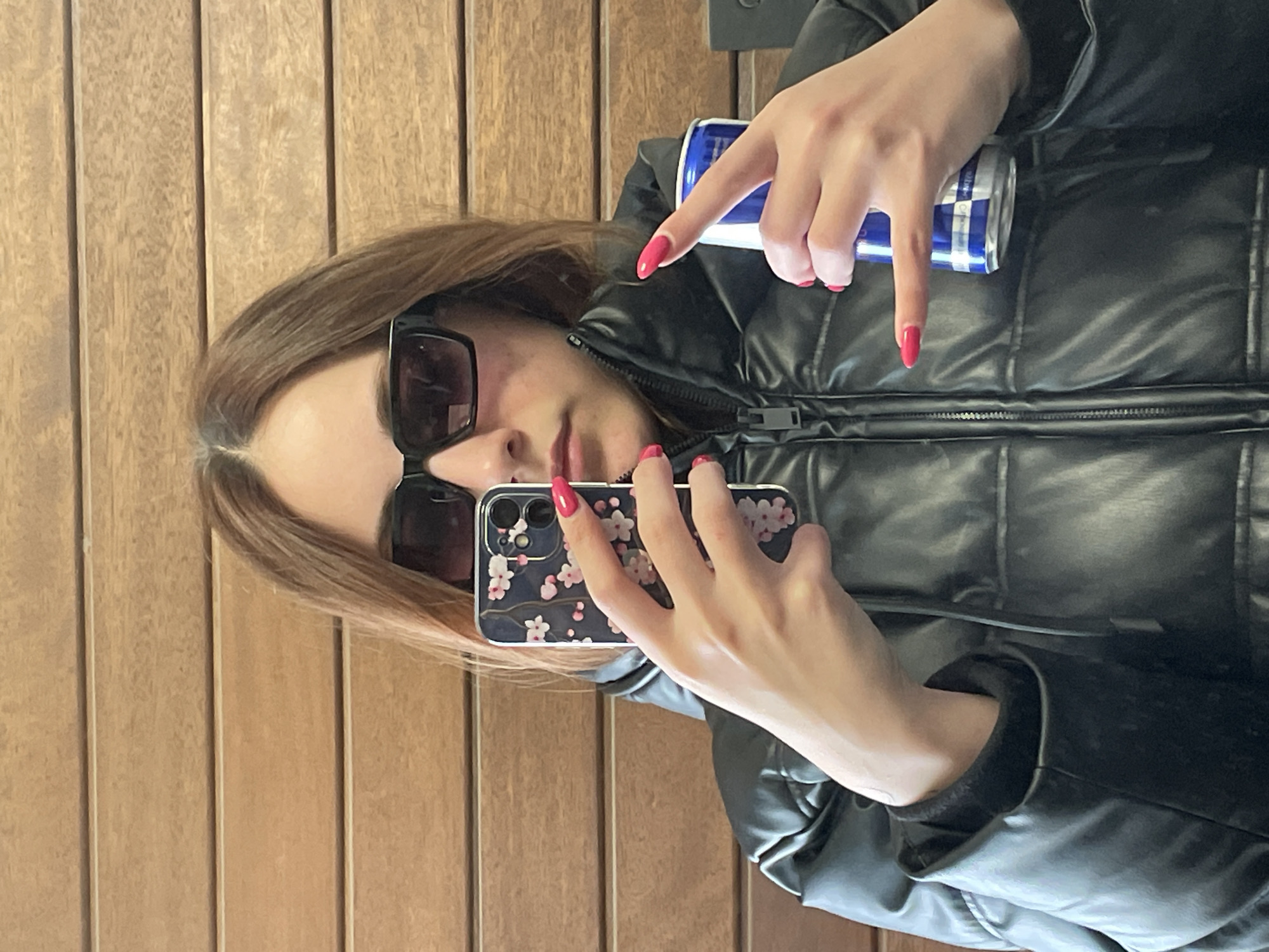 A woman is taking a selfie in front of a wood paneled wall. She is wearing a black puffy jacket and sunglasses. She is holding a phone in one hand, with a floral patterned case, and a blue and silver can in the other. Her nails are painted red. The location cannot be determined from this image.