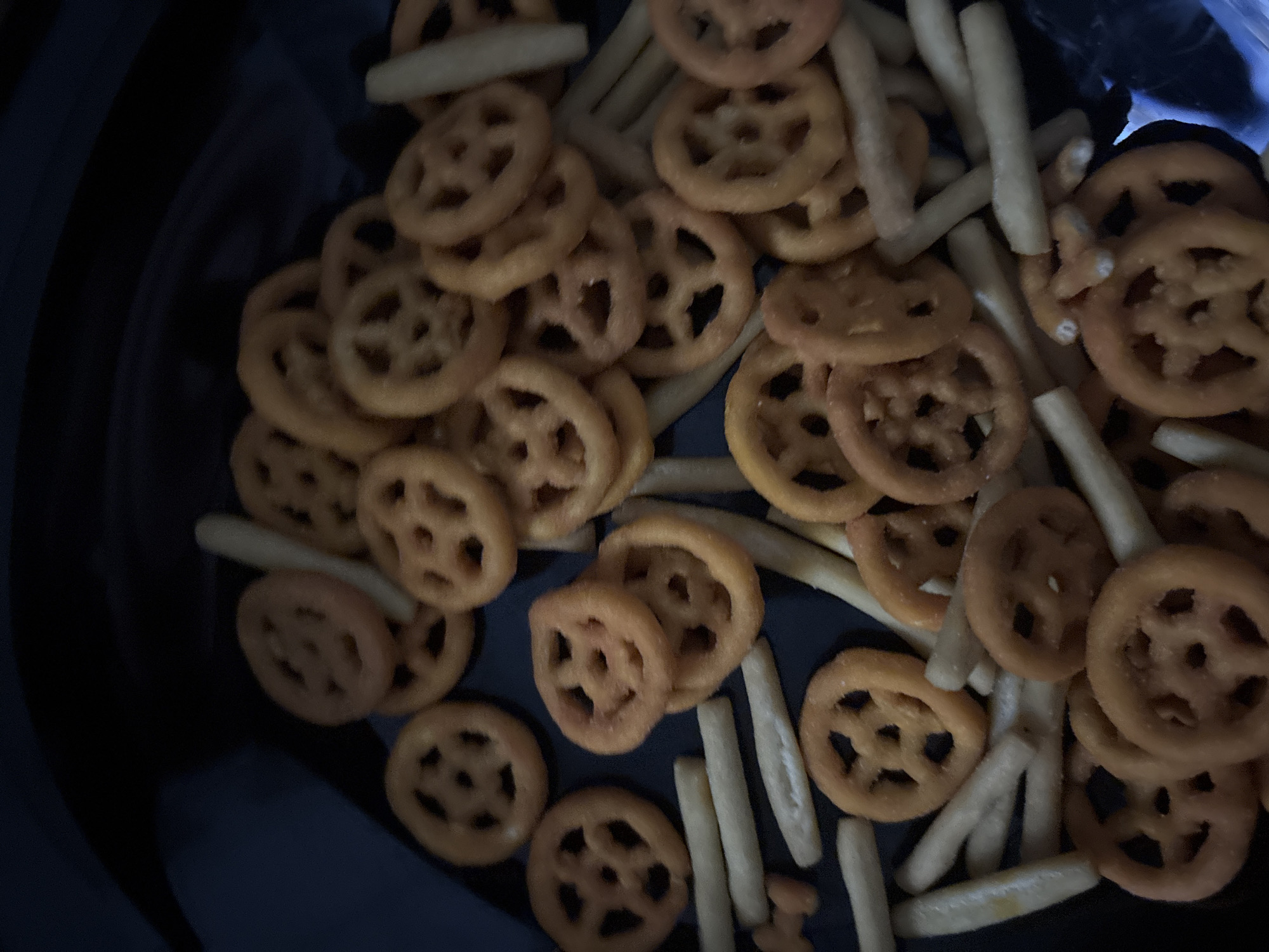 The image shows a close-up of a pile of wagon wheel-shaped pasta and skinny french fries-like sticks on a dark surface. The scene is devoid of any people. There is no discernible indication of its location in Buenos Aires, Argentina, or any other city. The objects are simply assorted snack foods. There are no evident actions or interactions taking place. The environment is limited to the surface the snacks are resting on.