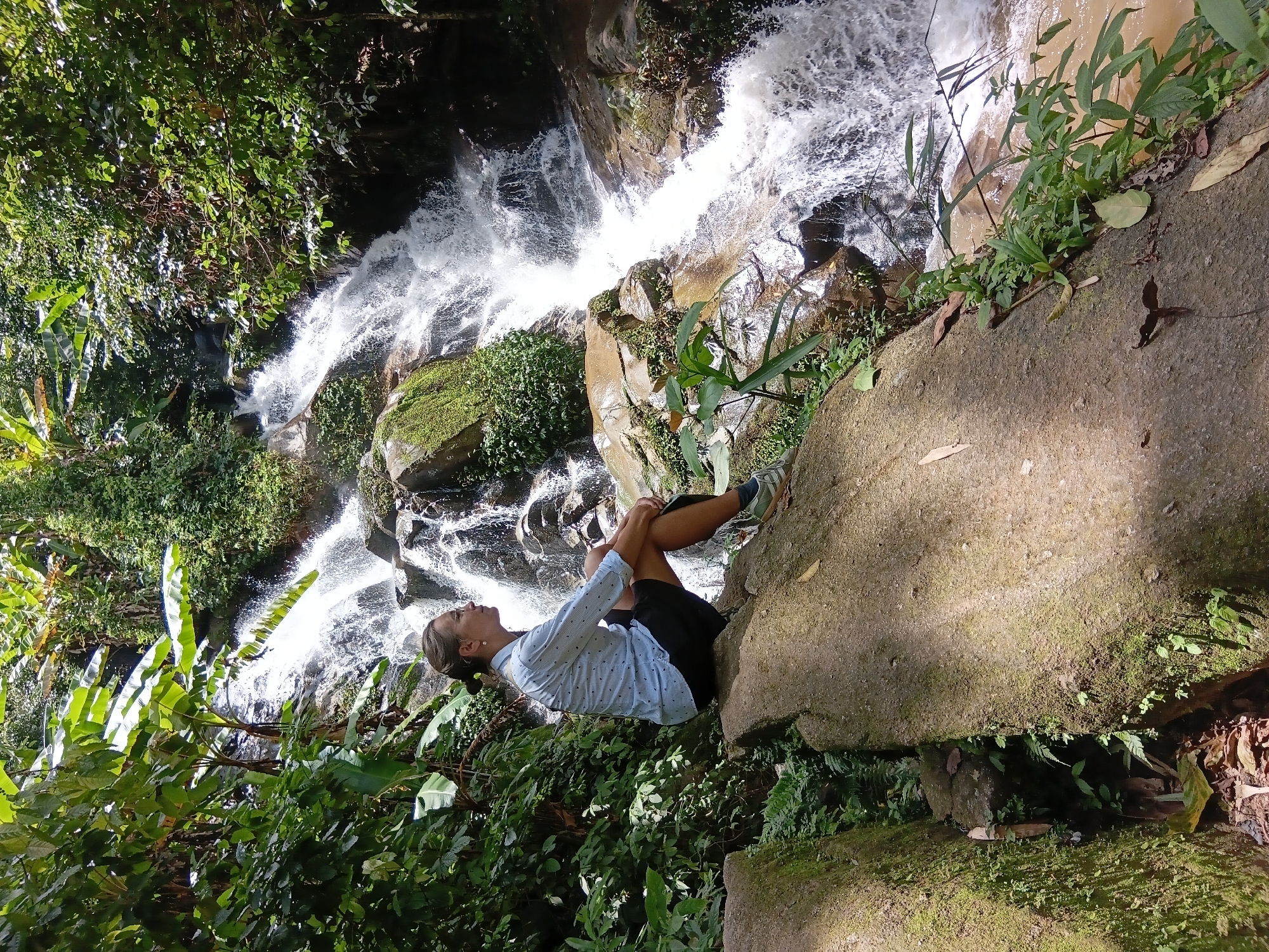 A woman with brown hair pulled back and wearing glasses sits on a large rock formation, facing left in profile. She is dressed in a light blue, long-sleeved button-down shirt with a small pattern and black shorts. Her legs are crossed at the ankles. She is positioned in the foreground on the left side of the image, with a waterfall cascading down rocks in the background. The waterfall is surrounded by lush green foliage, including large-leafed plants and trees, characteristic of a tropical environment. The water is white and frothy as it descends. The rock she is sitting on is large, textured, and appears damp, with moss or small plants growing on its surface in places. The overall scene depicts a natural landscape with the woman observing the waterfall. The location is identified as Ban Pong Nam Ron, Thailand.