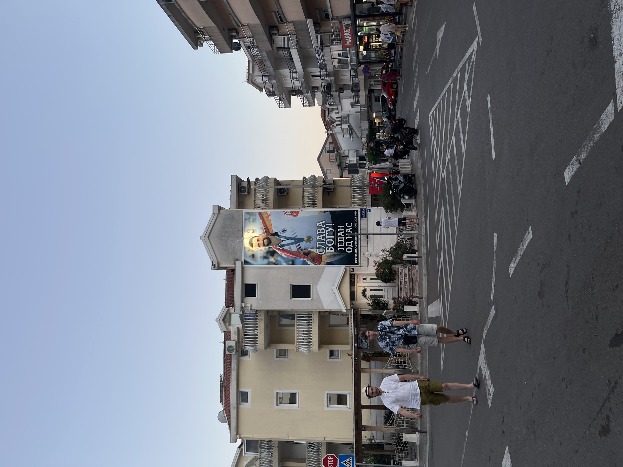 This is a full-shot, eye-level, outdoor photograph of a street scene in Budva, Montenegro. The image is captured during the daytime, with the sky being a clear, light blue. Dominating the background are multi-story buildings with balconies. A large banner is prominently displayed on the side of one building, featuring a man in athletic attire, possibly a medal winner, with text in a foreign language. In the middle ground, two men are walking on the street, dressed in casual summer clothing. The street itself has crosswalk markings. Several motorcycles are parked on the side of the road. In the right background, there is a market sign. The overall atmosphere appears to be that of a bustling urban area.
