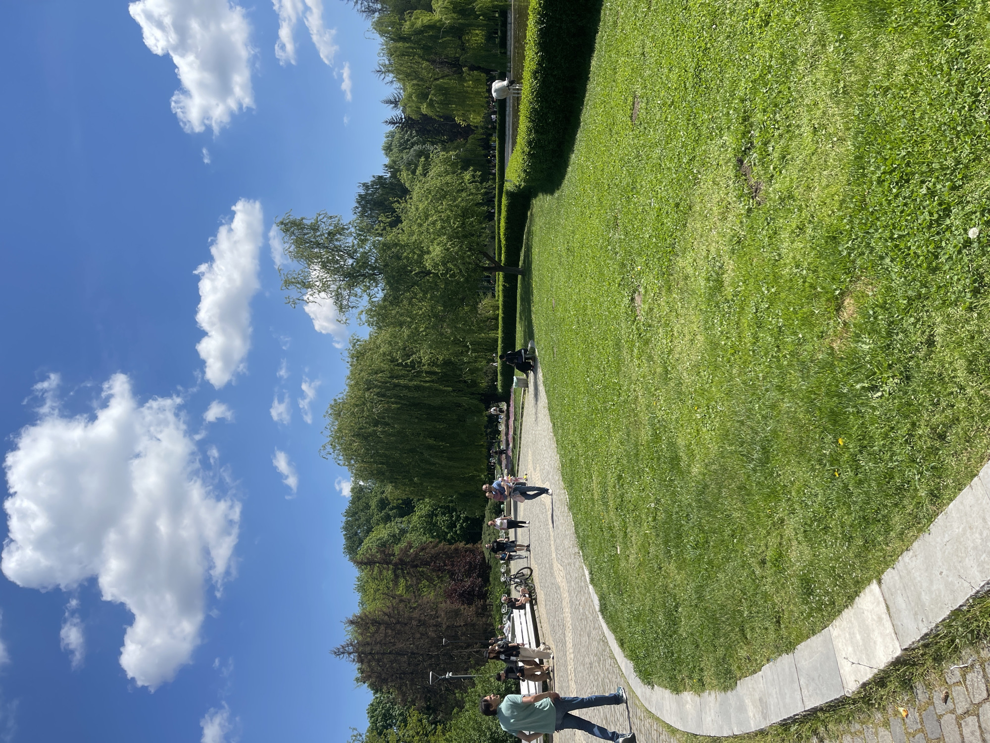 The image shows a park scene in Sofia, Bulgaria, likely Borisova Gradina Park, characterized by open green spaces and walkways.
The sky is blue with scattered white clouds. The foreground is dominated by a large, manicured lawn bordered by a stone pathway. Along the pathway are several benches where people are sitting and resting. A few people are walking along the pathway. The area is populated with various trees, including weeping willows. Beyond the trees, other greenery suggests the park extends further.