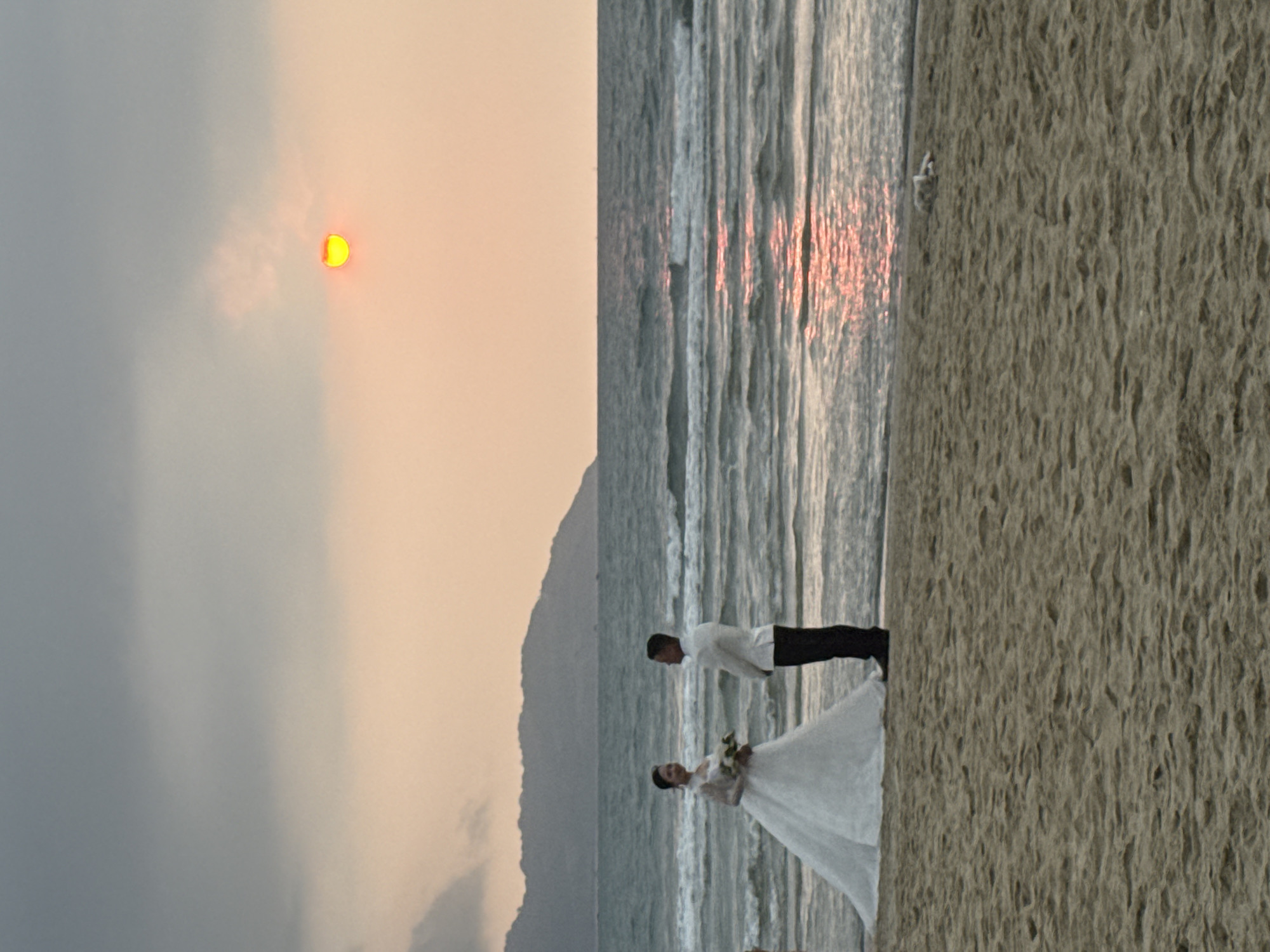 The image displays a serene beach scene, likely during sunset or sunrise, featuring a couple in wedding attire. The setting includes a sandy beach in the foreground, meeting the ocean with gentle waves. In the middle distance, the silhouette of mountains or hills is visible on the left side.
The main subjects are a bride and a groom. The bride, positioned on the left, is wearing a flowing white wedding gown and holding a bouquet of light-colored flowers. She is looking to her right. The groom, standing to the right of the bride, is dressed in a white long-sleeved shirt and dark trousers, appearing to look down or towards the bride. They are standing on the wet sand close to the water's edge.
A bright, distinct yellow sun, partially obscured by atmospheric haze or clouds, is prominent in the upper right portion of the sky. The sky transitions from a soft orange-yellow hue around the sun to a muted grey-blue further left. The ocean's surface reflects streaks of reddish-orange light from the sun, particularly visible in the waves. The overall atmosphere of the image is romantic and peaceful, suggesting a wedding photoshoot. The location context provided is Đà Nẵng, Vietnam. There is no visible text in the image.