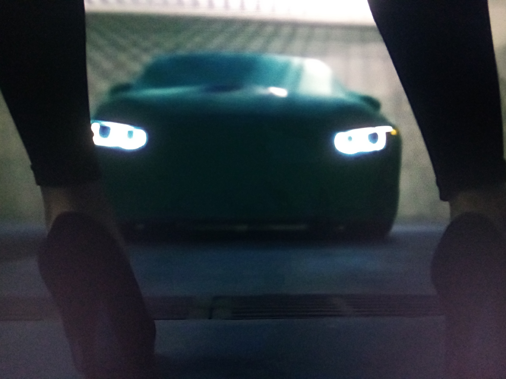 The image shows a dark green car with its headlights on, parked inside what appears to be a garage or covered parking area. The perspective is low, as if taken from the ground looking up at the car. The car's front is partially obscured by two figures, visible from the legs down, standing in the foreground. These figures are wearing dark pants and what look like shoes or lower legs of trousers. The scene is dimly lit, with the car's bright headlights illuminating the immediate area. The background shows a textured wall, possibly concrete or metal, with horizontal lines. The overall impression is one of waiting or preparing to depart. The location context of Chirakchi, Uzbekistan, suggests this is likely a private or public parking space in that region. No text is visible in the image.