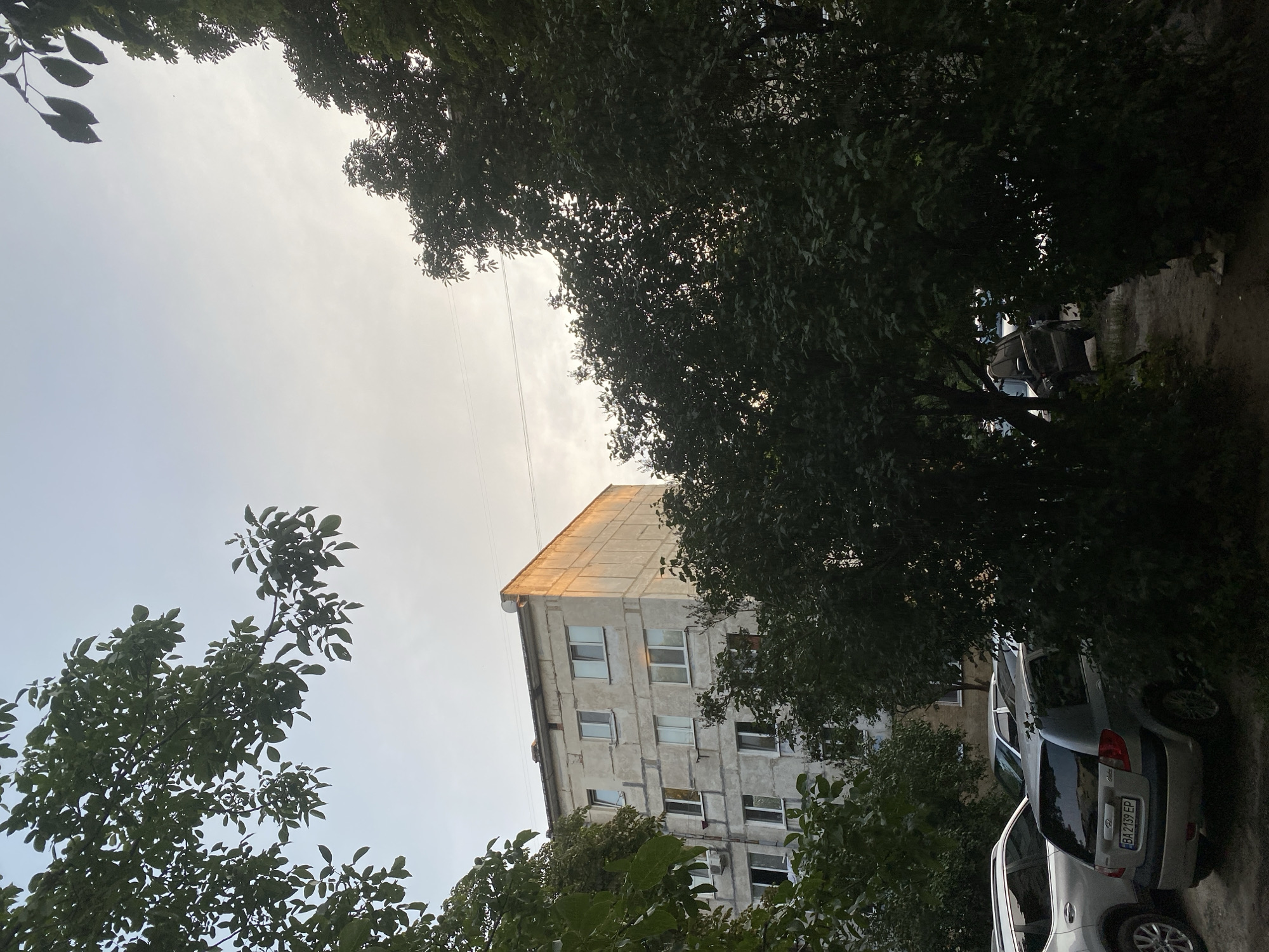 This is a photograph of an overcast sky above a residential area in Poltava, Ukraine. In the foreground, there are trees with lush green foliage partially obscuring the view. To the left, the side of a silver SUV is visible. Further to the left, a lighter colored vehicle is parked. In the center of the image, a silver hatchback car is parked. Behind the vehicles, a concrete building with multiple stories and numerous windows stands. The side of the building is illuminated by the setting sun, casting an orange hue. Power lines are visible crossing the sky.