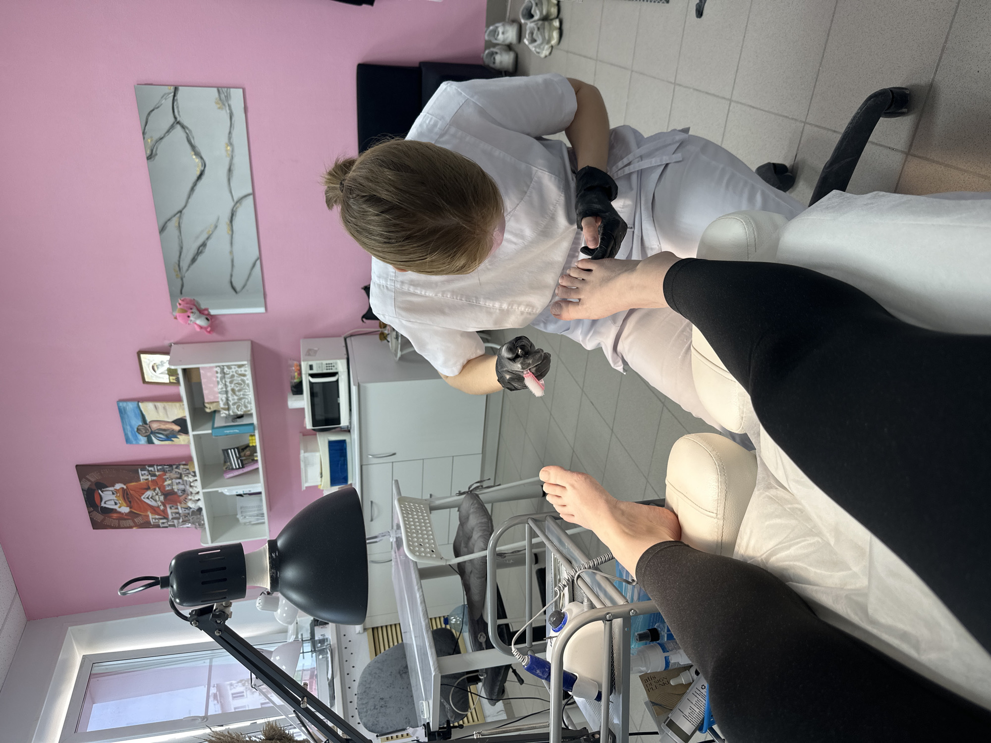 A woman is providing a pedicure to a client in a salon. The salon has pink walls and tiled floors. The client is wearing black leggings and is reclined in a white chair. The pedicurist is wearing a white uniform and black gloves. She is holding a small brush and is focused on the client's feet. A black lamp illuminates the area. A microwave, shelves with objects, and a painting are visible in the background. The location is Chaikovsky, Russia.