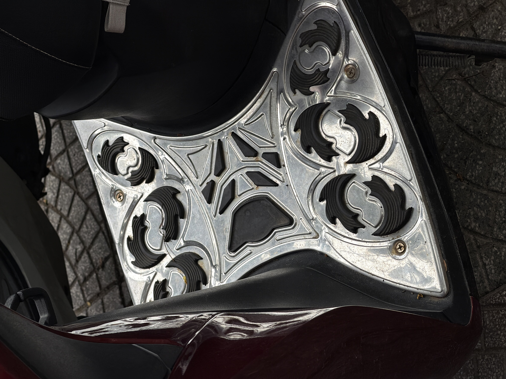 The image shows a close-up view of the footrest area of a scooter or motorcycle. The footrest is made of patterned metal with a design consisting of repeating swirl shapes and a central geometric pattern. The surface appears to be secured with screws. Parts of the seat, tire, and body of the vehicle are also visible. The setting is an outdoor environment with pavement. There are no visible people or actions captured in the frame. While the prompt suggested the location is Đà Nẵng, Vietnam, this cannot be verified from the image itself.
