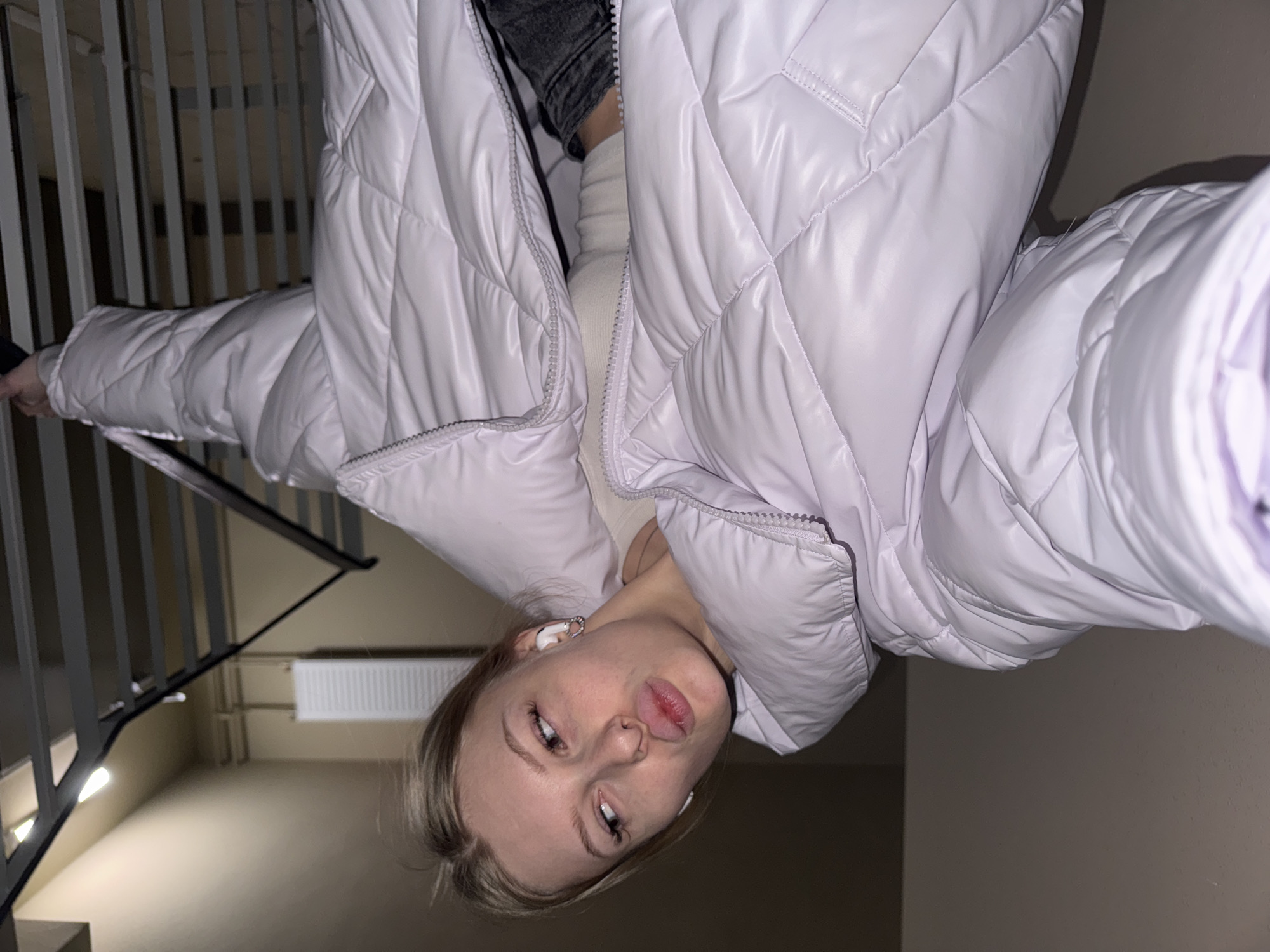 A young woman is featured in an indoor setting, likely a stairwell or hallway in Perm, Russia. The image is oriented upside down, captured from a low angle looking upwards towards the camera. She has blonde hair and fair skin, with slightly parted, full lips. She is dressed in a light, possibly lavender or white, quilted puffer jacket worn over a light-colored turtleneck or top, paired with dark pants or jeans. White wireless earbuds are visible in both of her ears.
The background shows a black metal staircase railing on the left, and a white wall-mounted radiator on the right against a plain, light-colored wall. The scene is illuminated by artificial indoor lighting. The woman maintains a neutral expression, gazing directly at the camera. No text is visible in the image.