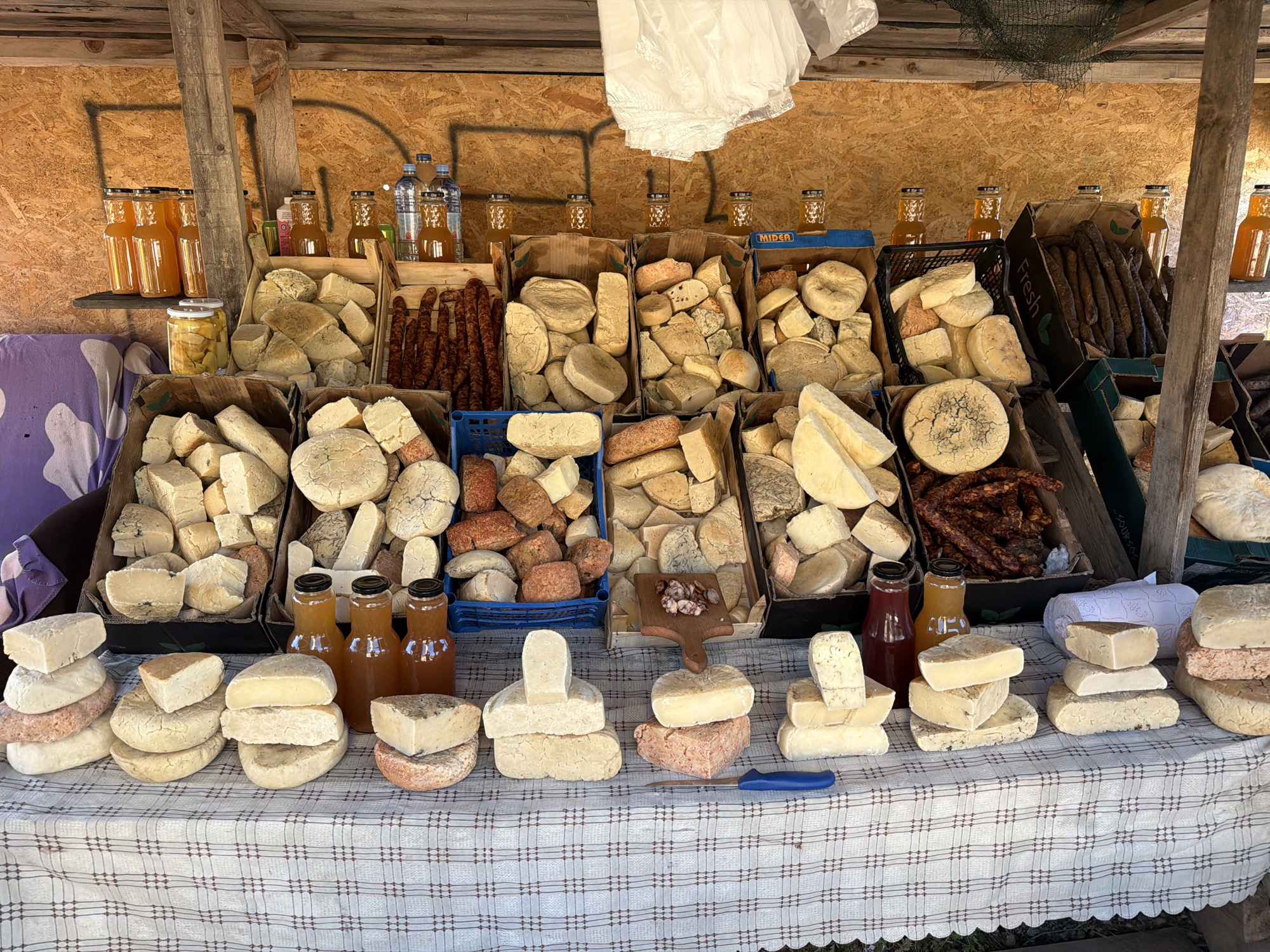 At a market stall in Ponikvica, Montenegro, a variety of cheeses and bottled beverages are displayed for sale. The stall features wooden shelves and panels, with a checkered tablecloth covering the display surface. Large crates overflow with numerous pieces of cheese of various shapes, sizes, and textures, some appearing aged and crumbly, others smooth and firm. Several boxes are filled with dark, cured sausages. On the shelves above, rows of clear glass bottles containing amber-colored liquids, likely juices or preserves, are arranged. Water bottles and more juice bottles are also visible. In the foreground, stacks of cheese are arranged on the tablecloth, with a blue-handled knife resting on the fabric. The overall scene suggests a local market offering artisanal food products.