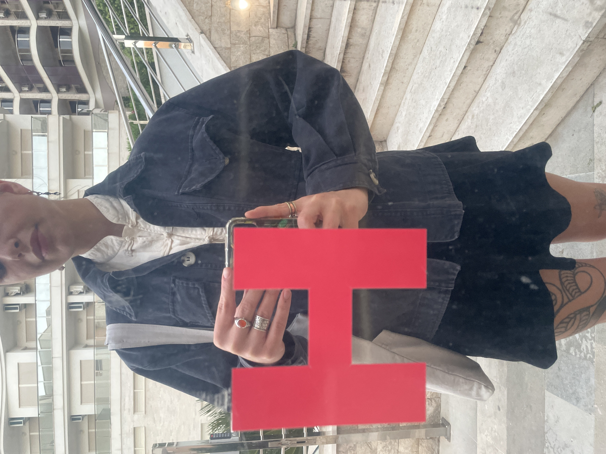 The image depicts a reflection in a window or mirrored surface, showcasing a person wearing a dark denim jacket over a white collared shirt and a black skirt. The person has short hair and is wearing rings on their fingers. A light-colored bag is slung over their shoulder. The background visible through the reflection includes a modern building with repeating window patterns and a staircase. The setting is Boreti, Montenegro, as evidenced by the architectural style of the building and the typical landscape. The person appears to be taking a photo or looking at their phone, which is partially obscured by a red, geometric shape on the window. A glimpse of a tattoo on the person's leg is visible. The reflection also shows parts of a stone staircase.