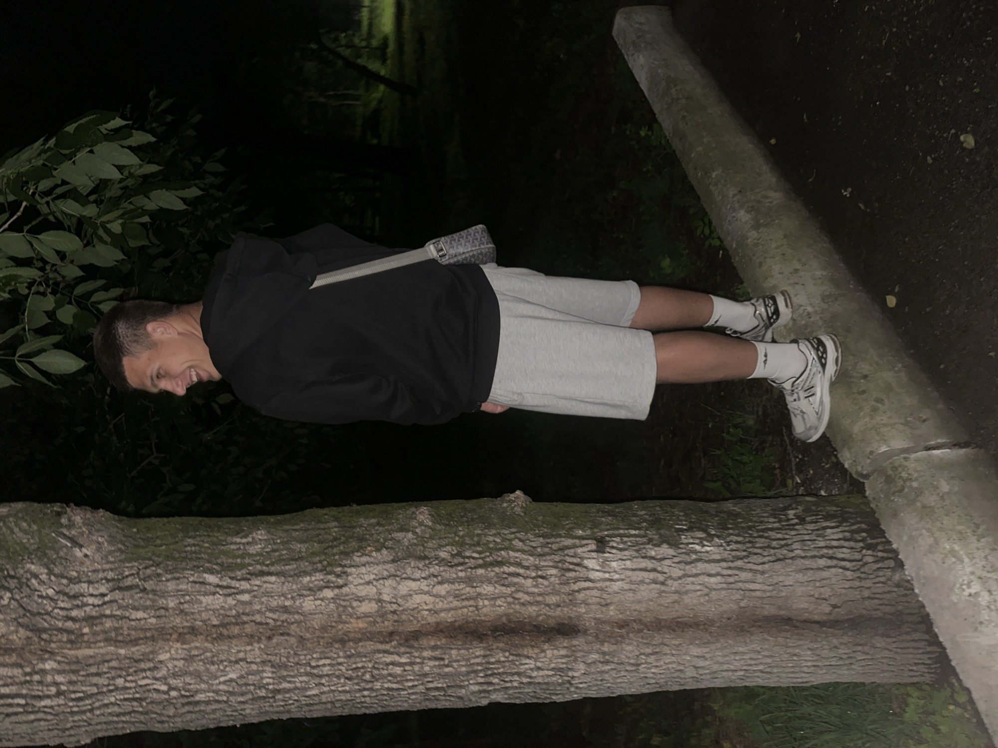 A young man is standing on a concrete curb in a park at night. He is wearing a black hoodie, grey shorts, white socks, and athletic shoes. He has a small messenger bag slung over his shoulder. He is smiling and looking over his shoulder at the camera. The background is dark, with trees and foliage visible. There is a large tree trunk to the left of the man. The ground is paved. The scene appears to be in a park, possibly in the city of Birobidzhan, Russia, given the context.