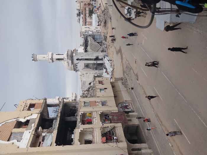 The image depicts a wide street in Khan Yunis, Palestinian Territory, under an overcast sky. On the left, a multi-story building exhibits extensive structural damage, with several floors exposing collapsed sections, shattered windows, and rebar. Its facade is pockmarked with numerous small impact holes. Laundry, including red and yellow items and a patterned rug, hangs from some balconies.
Across the street, a mosque with a prominent, light-colored minaret and dome stands amidst widespread destruction. Its lower sections and surrounding structures are largely reduced to rubble, while the minaret itself shows signs of damage. Extensive piles of building debris and collapsed materials are scattered between the street and the mosque.
The paved street, marked with white lane lines, is occupied by approximately two dozen individuals, including adults and children, seen walking, running, and congregating in small groups. A white sedan, appearing damaged, is visible on the far right side of the street. Partial view of a utility pole with cables is present in the extreme right foreground.