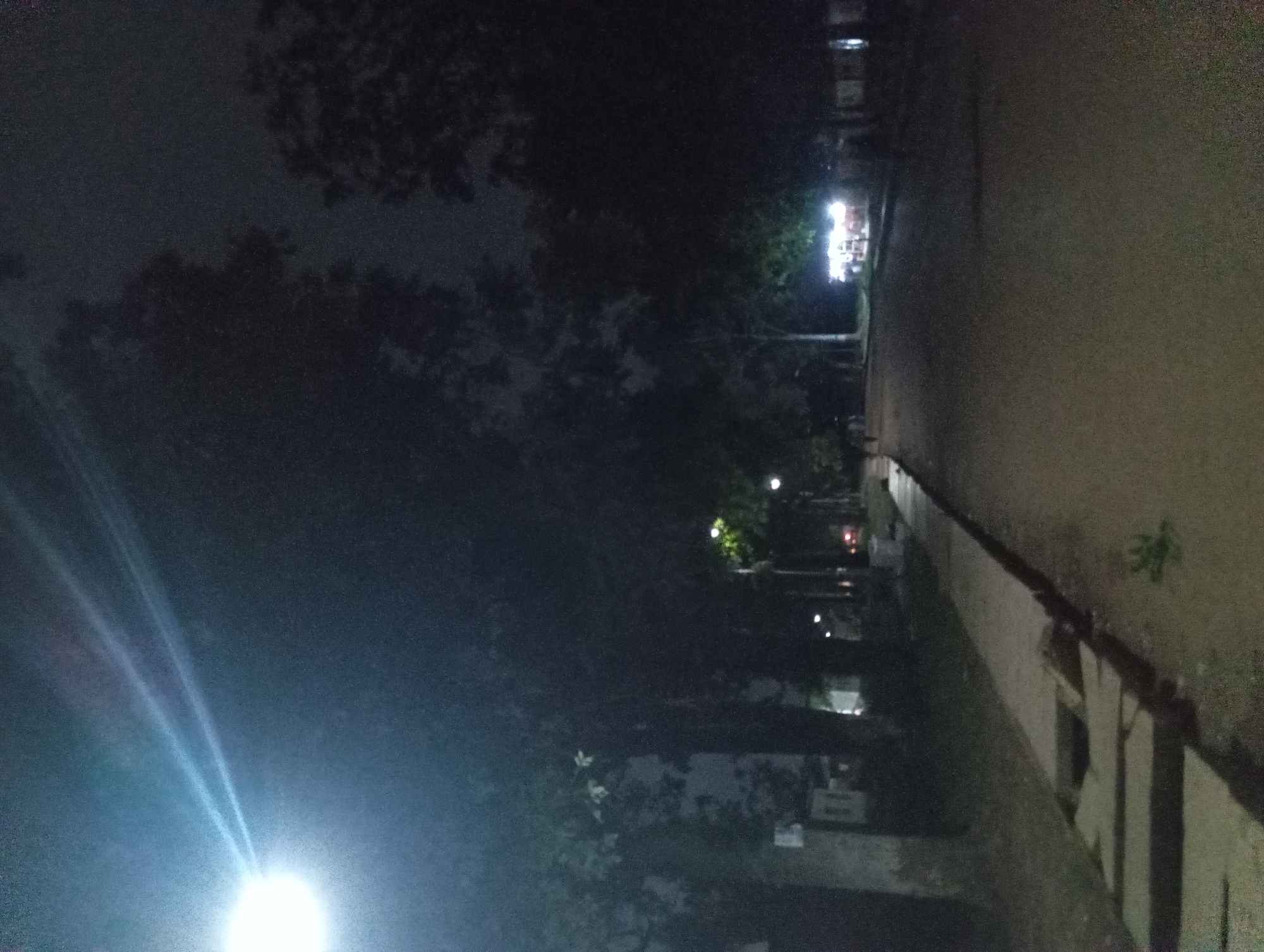 This is a nighttime photograph taken from a low angle, looking down a paved path or road. A bright overhead light casts strong beams across the upper left of the image, illuminating a portion of the dark, cloudy sky. Silhouetted trees line the sides of the path, with some foliage visible against the dimmer sky. In the mid-ground, a small structure or food cart with bright lights is visible on the right side of the path, suggesting a commercial area or event. A few figures are faintly visible in the distance, walking along the path. The path itself is lined with a raised concrete border, and in the foreground, there are some broken concrete slabs or steps. The overall impression is of a quiet, possibly urban park or street scene at night.