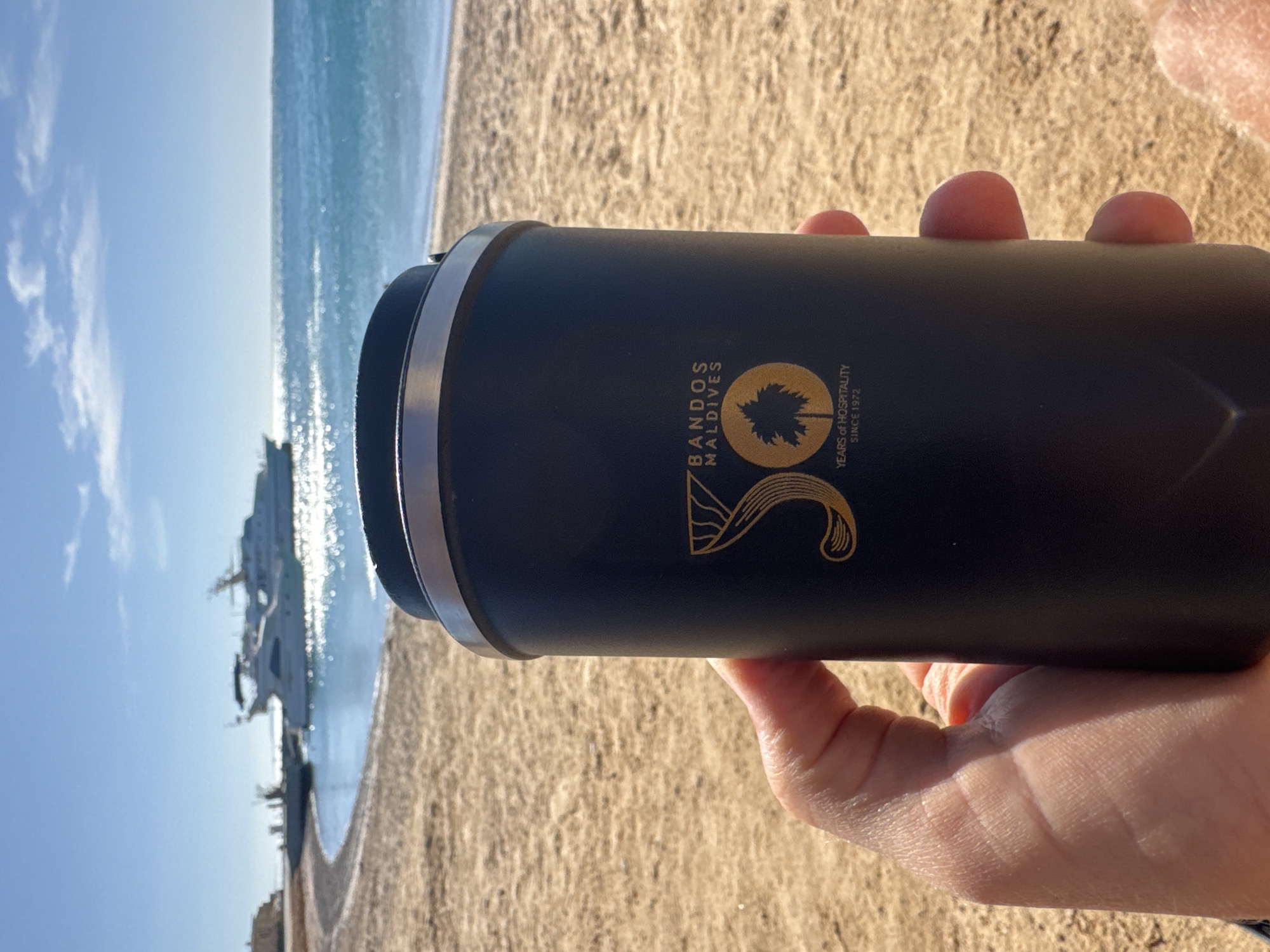 The image shows a person holding a black cylindrical container with silver trim. The container has a gold logo that reads "BANDOS MALDIVES" and "YEARS OF HOSPITALITY SINCE 1972". The person is holding the container with their right hand, with fingers visible on the top. In the background, there is a beach with light brown sand, turquoise water, and a blue sky with some clouds. A structure resembling a building or dock is partially visible in the water. The location is likely a beach at Bandos Maldives, though the specific city is unknown.