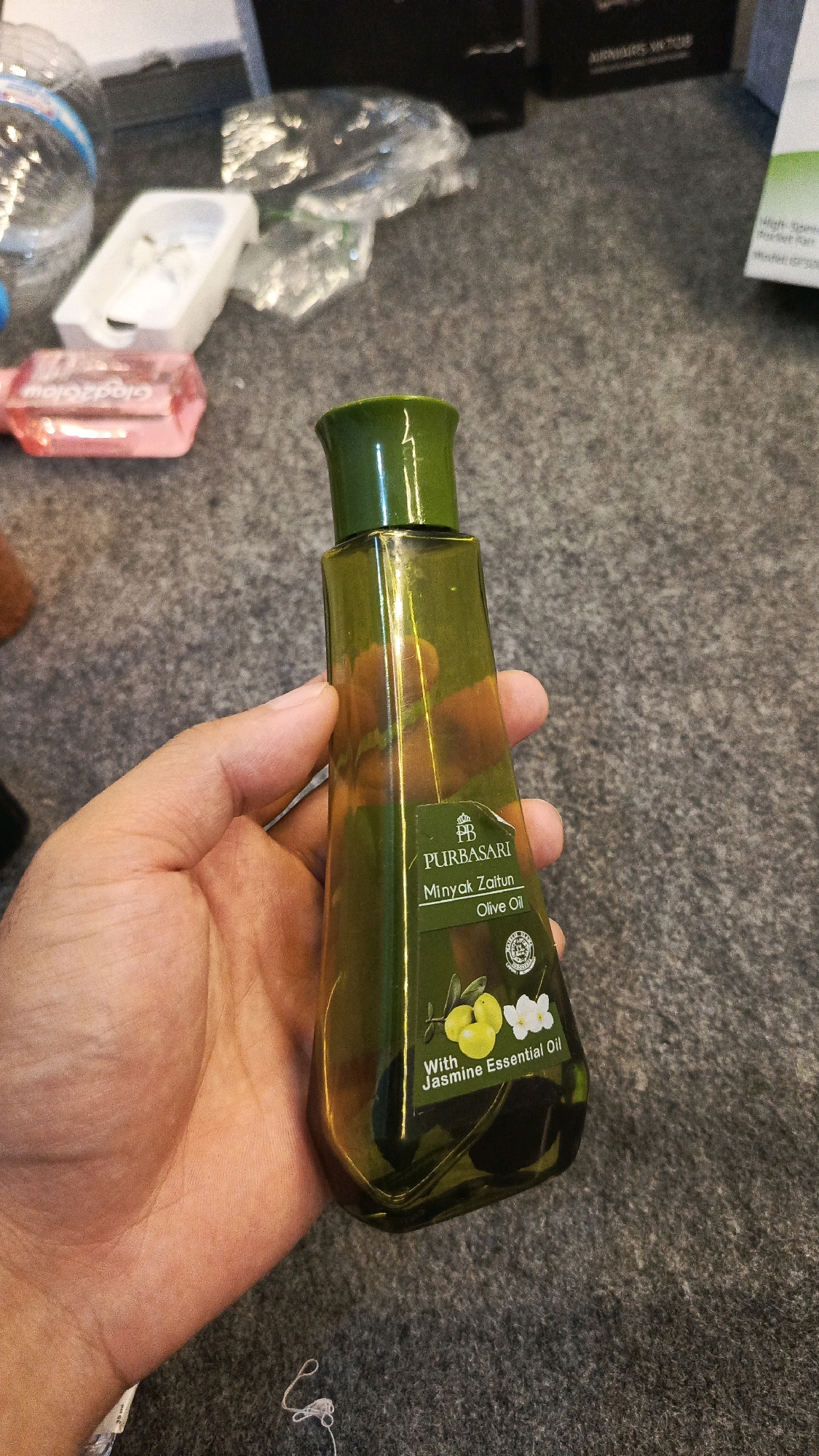 A hand holds a green glass bottle of Purbasari brand olive oil. The bottle is rectangular with a green cap and a label that reads "PURBASARI Minyak Zaitun Olive Oil With Jasmine Essential Oil." The bottle is slightly angled, and the hand is holding it from the side, obscuring part of the bottle's surface. In the background, the scene appears to be an indoor setting with a gray, textured surface, possibly carpet or fabric. There are other items in the background, including plastic packaging and what looks like a black electronic device. The lighting is moderate, casting some reflections on the bottle. The image does not provide any information about the specific location within the city of Babadan, Indonesia.