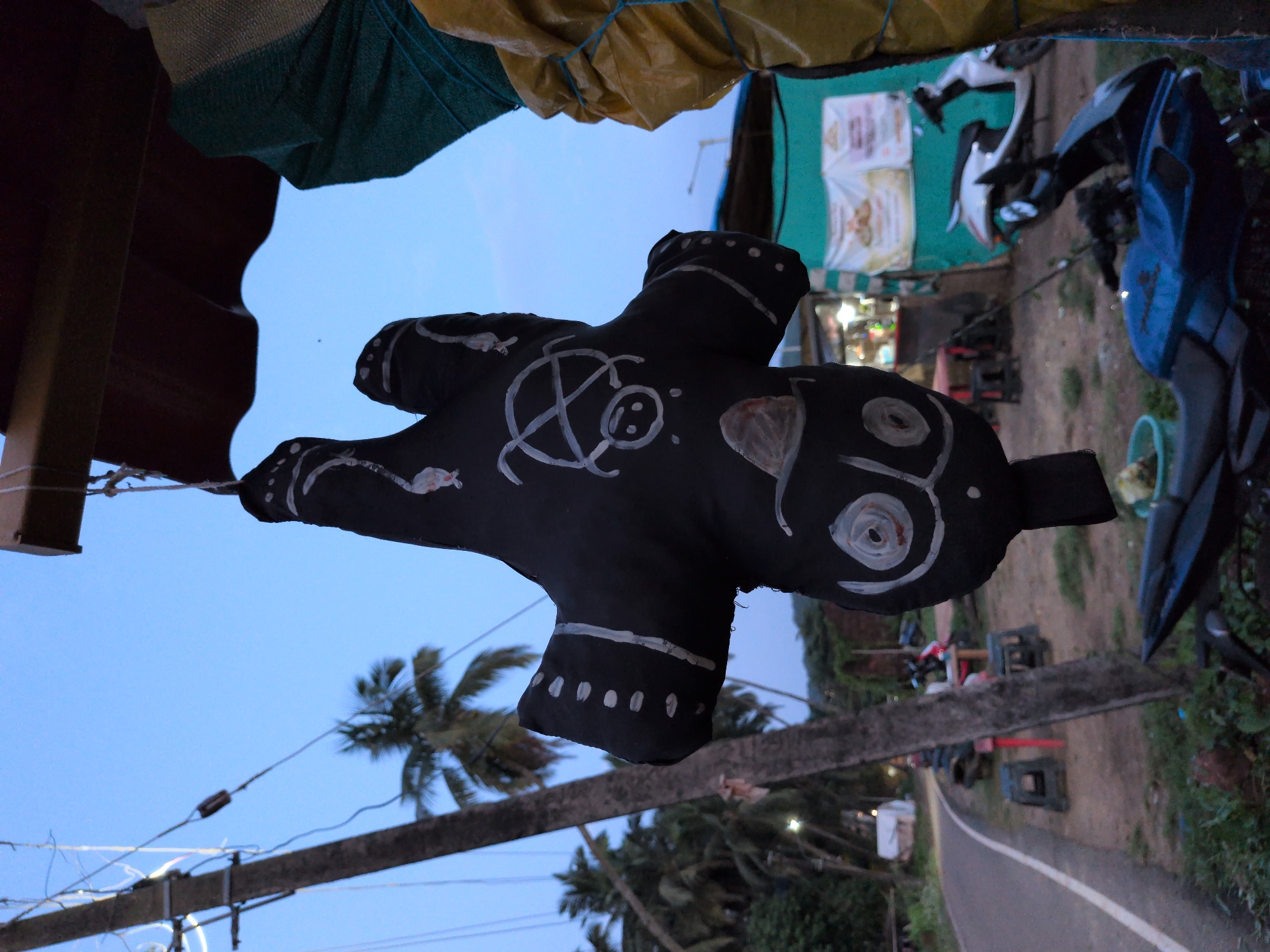 In Badem, India, an upside-down black effigy with white drawings hangs from a wooden structure, likely at a roadside stall. The effigy's design includes circular patterns resembling eyes and a cross-like symbol with a face. Behind it, a road curves to the left, bordered by palm trees. Several motorcycles are parked nearby, and a small shop with signs is visible in the background. The sky is a clear blue, suggesting it is daytime.