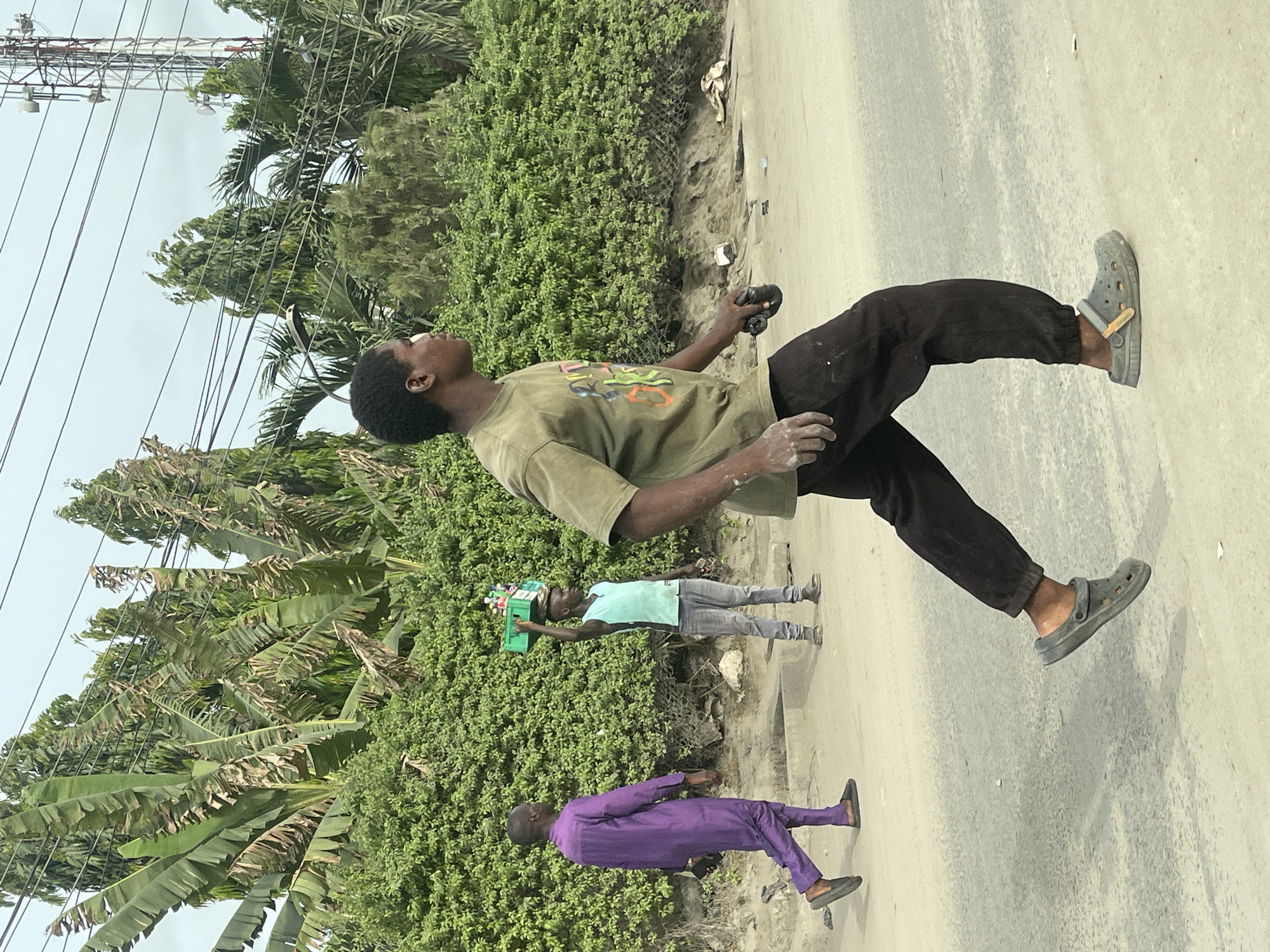 The image captures an outdoor street scene in Lekki, Nigeria, under an overcast sky, likely during the day. The primary subject, a young man, stands in the foreground, facing right but looking upwards intently. He wears an olive green t-shirt with a vibrant, abstract graphic, dark pants, and dark gray Crocs. His hands are noticeably smudged with a white substance, possibly dust or paint, and he holds a dark, cylindrical object in his right hand.
To the left of the main subject, another person, possibly a child or young teenager, walks rightward, balancing a green plastic crate filled with small bottles (presumably drinks) on their head, typical of a street vendor. Further behind them, a man dressed in a bright purple traditional Nigerian outfit (possibly an agbada) and dark shoes also walks towards the right side of the frame.
The setting features a mix of natural and man-made elements. A light-colored road or dirt path occupies the right side of the frame. The left side is dominated by lush green vegetation, including dense bushes, several large banana trees with broad leaves, and taller palm trees. In the upper left background, a large metal structure, possibly a telecommunications mast or electricity pylon, is visible with numerous power lines stretching across the sky. The overall scene depicts everyday activity along a roadside in a semi-urban or suburban area of Lekki. No explicit readable text is visible in the image, other than the abstract design on the main subject's shirt.