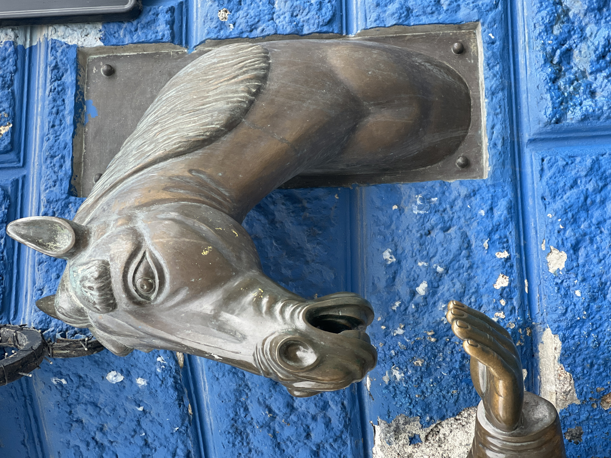 The image shows a bronze sculpture of a horse's head protruding from a textured, bright blue wall. The horse's mouth is open, with a spout-like structure inside, suggesting it might be a water fountain or a drainpipe. Below the horse's head, a bronze sculpture of an outstretched hand is visible, with its palm facing upwards, as if to catch water or receive something. The wall appears to be made of brick or plaster, with visible imperfections and some peeling paint. The overall aesthetic suggests an artistic or decorative element on a building, likely in an urban setting. This sculpture is located in the city of Yekaterinburg, Russia, specifically on the wall of the building at 48 Komsomolskaya Street, serving as a public water fountain.
