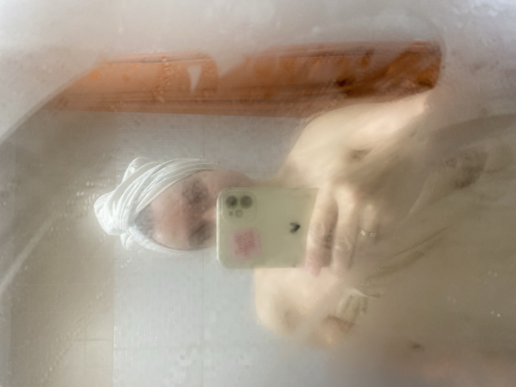 The image is a selfie taken in a bathroom, likely inside a shower stall or behind a steamy mirror. A person with a towel wrapped around their head is holding a phone, presumably to take the picture. The phone has a light-colored case and a sticker on the back. The background is obscured by condensation, but hints of a wooden shelf or fixture are visible. There is no verifiable evidence to support the image's location within the city of Chaikovsky, Russia.