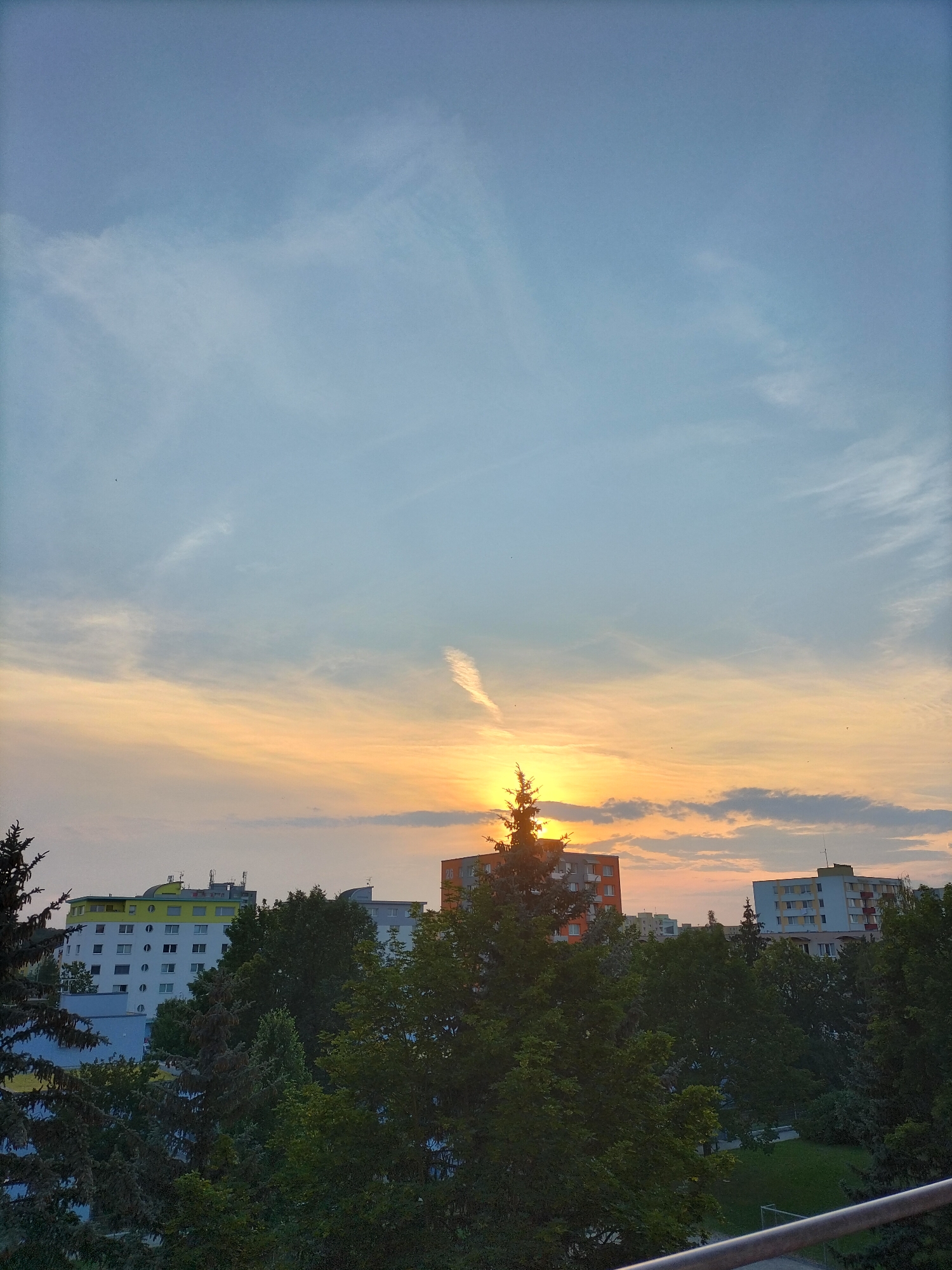 This is a vertical, wide-angle photograph capturing a sunset over an urban residential area in České Budějovice, Czechia. The sky dominates the upper two-thirds of the image, displaying a gradient of soft blues and pale yellows, indicative of either dawn or dusk. Wispy clouds streak across the sky, with a prominent contrail visible near the setting sun. The sun itself is partially obscured by a tall evergreen tree, casting a warm, orange glow across the horizon and illuminating the clouds.
Below the sky, a line of apartment buildings is visible, interspersed with lush green trees. The buildings vary in architectural style and color; one prominent building has a bright yellow band across its upper floors, while another is accented with orange and red. The trees in the foreground are dense and verdant, suggesting a temperate climate and the warmer months of the year. The overall scene evokes a sense of peace and tranquility, typical of a quiet evening in a residential neighborhood. The lighting suggests it is late afternoon or early evening. There is no visible text in the image.