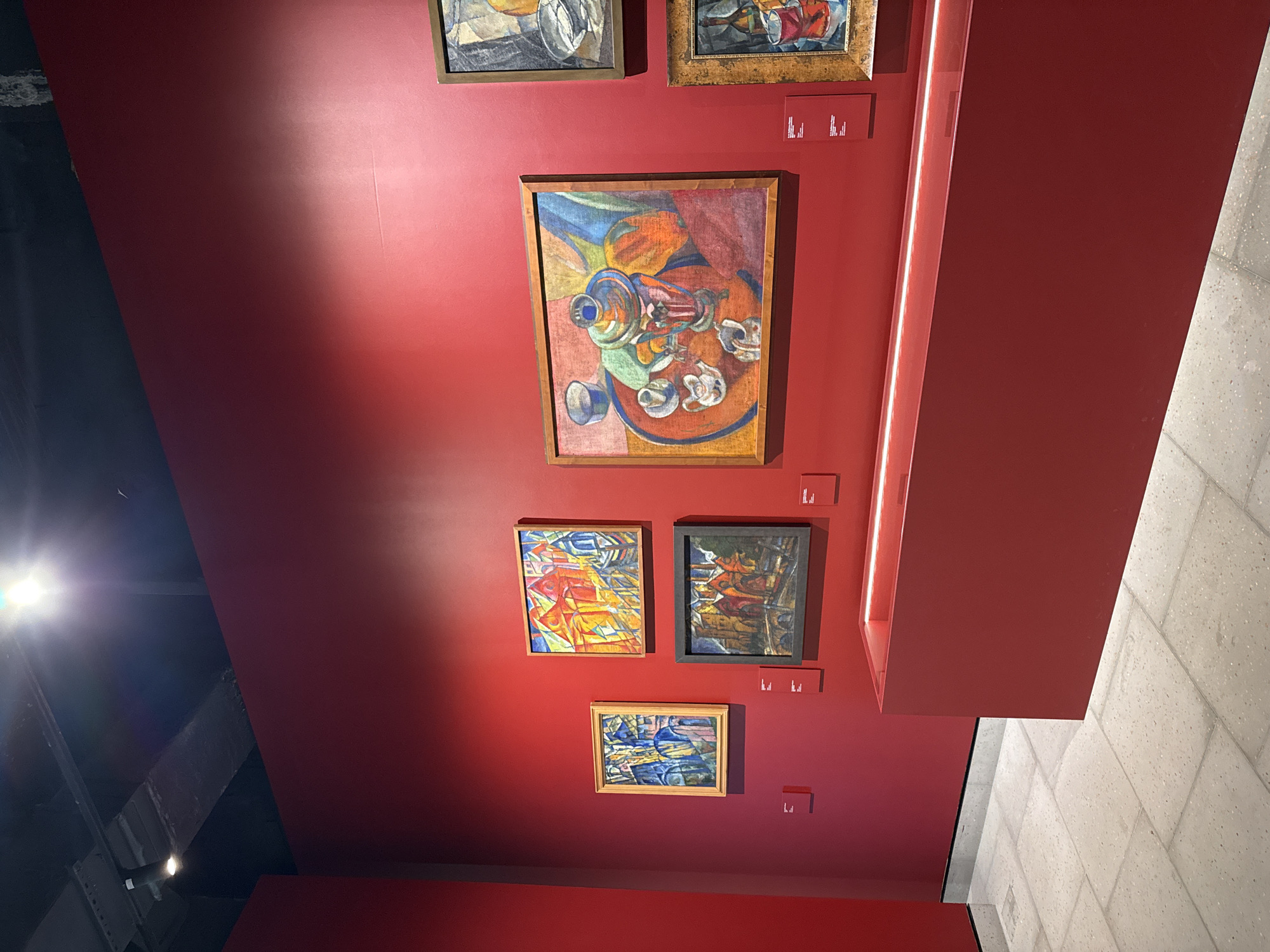 The image depicts a collection of framed paintings displayed on a red wall, likely within an art gallery. The paintings vary in style and subject matter, including still life arrangements and abstract compositions. Small rectangular plaques are positioned beneath each artwork, presumably containing information about the artist and title. The wall is lit by a spotlight in the upper left and a linear light fixture to the right. The wall is located in an art gallery in Perm, Russia. The floor is covered in light-colored stone tiles.