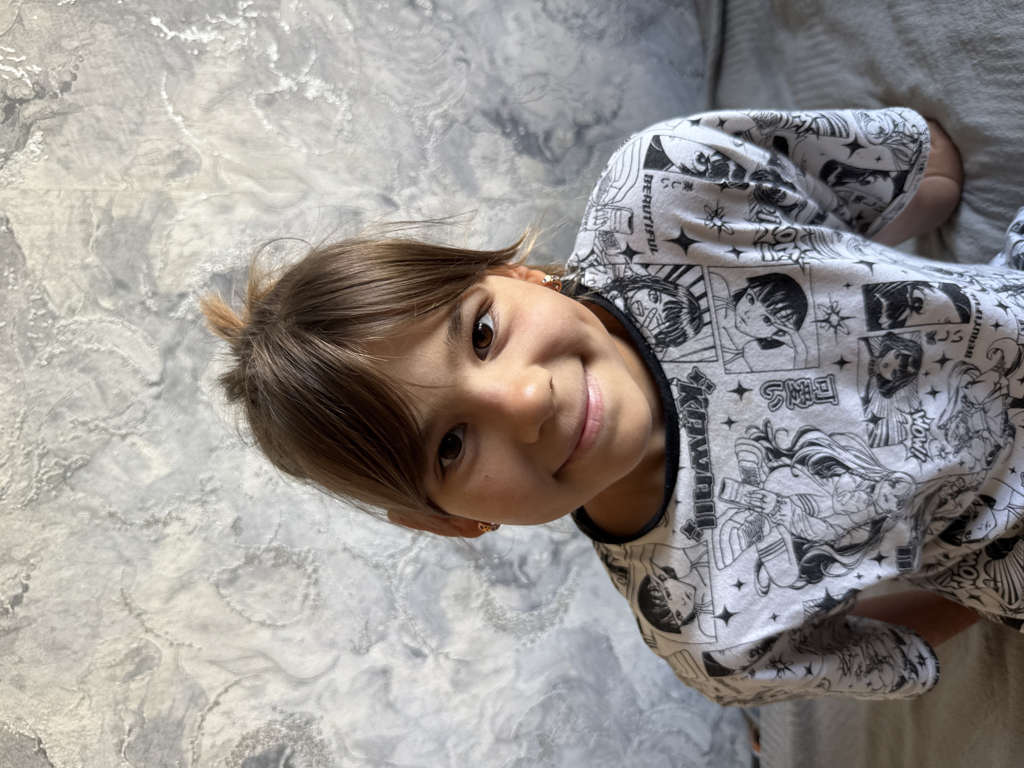 In Rusanovo, Russia, a young girl with brown hair tied up in a messy bun and wearing a t-shirt with a black anime-inspired print is captured in a medium shot. She has brown eyes, a slight smile, and a dimple on her right cheek. The background features a textured gray and white wall. The lighting suggests an indoor setting, possibly a room in a residence.