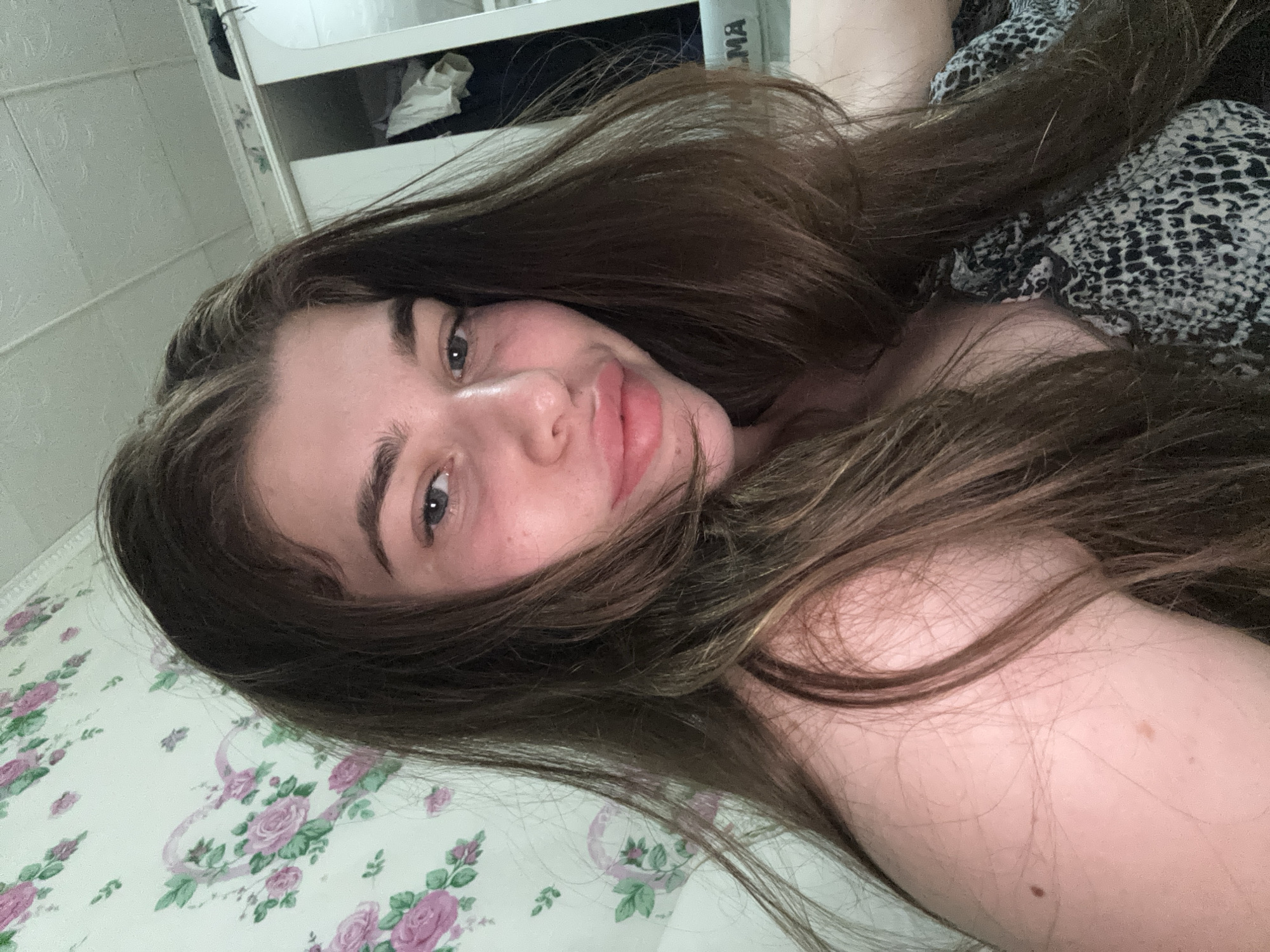 A young woman with long, brown hair is captured in a close-up, selfie-style photograph. She is looking directly at the camera with a slight smile. Her hair is loose and falls around her face and shoulders. She is wearing a patterned top. The background appears to be a room, with floral wallpaper visible on the left side and part of a closet or wardrobe on the right. The lighting suggests it may be indoors. The image does not provide any specific details that would allow for identification of the location within the city of Мещёры, Russia.