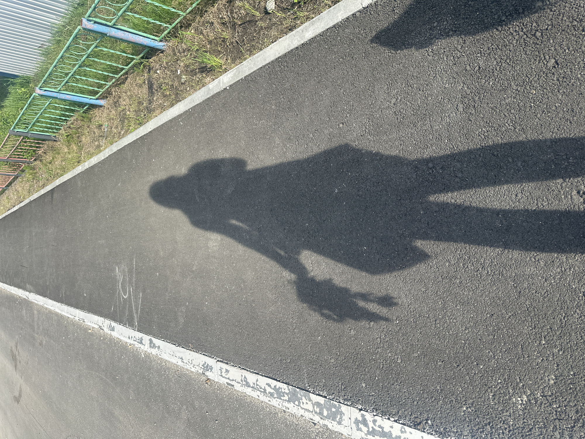 An outdoor scene depicts a paved asphalt pathway under clear daylight, with elongated shadows indicating either morning or late afternoon. The prominent shadow of a person, presumably the photographer, is cast centrally on the dark, textured asphalt, extending from the bottom foreground. The shadow's left arm appears to be holding the distinct shadow of a smaller, humanoid figure, resembling a doll or a young child.
To the left of the asphalt path, a weathered white line or curb separates it from an adjacent, lighter grey paved surface. On the right, a clean concrete curb borders a strip of dry, sparse green grass and dirt. Parallel to the path, a metal fence with decorative loops is installed; its sections are painted in faded green (with blue posts) and orange/red hues. Behind the fence, a portion of a corrugated light-colored metal wall is visible. Faint white chalk markings are present on the asphalt path near the center-left. A partial shadow of an additional figure or object is visible in the bottom right corner of the frame.