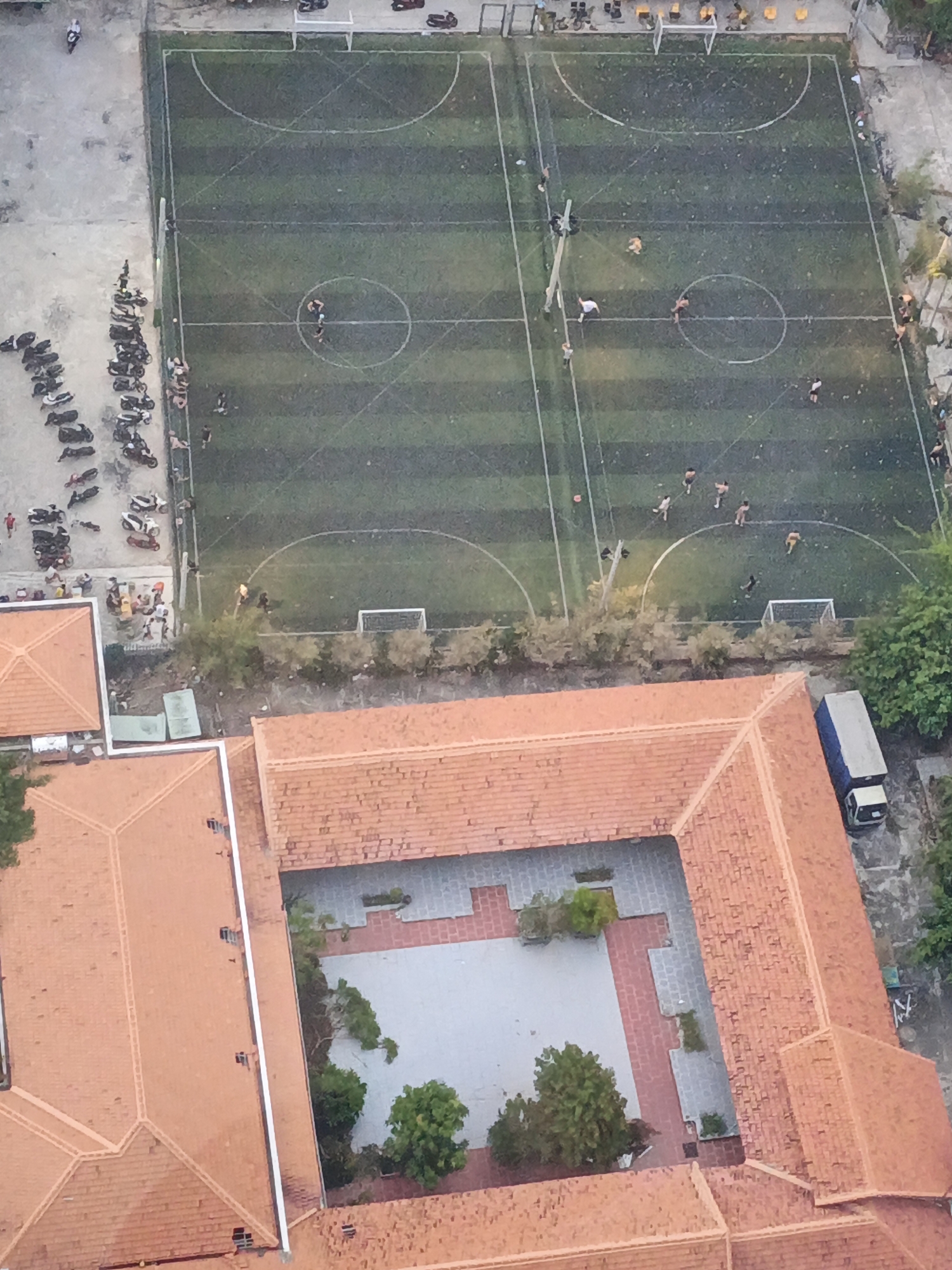 This aerial image captures a scene in Bac Nha Trang Ward, Vietnam, likely during daylight. The primary focus is a small, artificial turf soccer field with visible markings, including a center circle and semi-circles at each end. Several individuals, presumably playing soccer, are scattered across the field.
To the left of the soccer field, a long line of parked motorcycles and scooters is visible, suggesting a gathering or event nearby. A few people are standing or sitting near the motorcycles.
In the foreground and lower portion of the image, the terracotta-tiled roofs of a building complex are prominent. This building appears to have a central courtyard with paved areas and some greenery. A white van or truck is parked next to the building.
The overall impression is a snapshot of everyday life in a Vietnamese urban or semi-urban environment, combining sports, transportation, and architectural elements. The time of day appears to be daytime, and there are no immediate indicators of specific weather conditions beyond the general lighting. No visible text is present in the image.