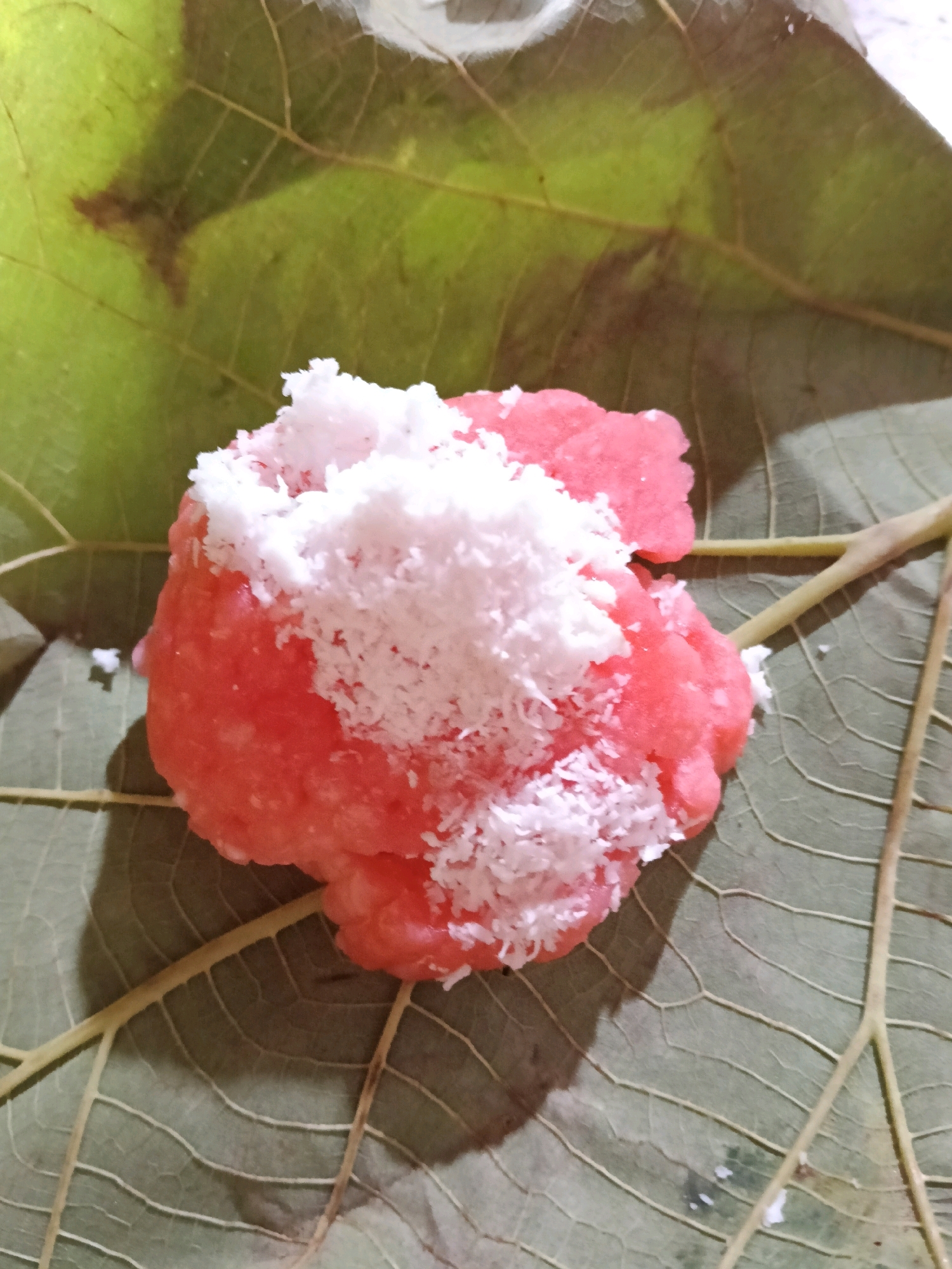 A close-up, overhead shot shows a pink, mound-shaped confection topped with shredded white coconut. The food item rests on a large, dried green leaf, which has visible veins. The leaf, in turn, is placed on a surface that casts a soft shadow. The scene appears to be set outdoors, possibly in a rural area, given the natural elements. The specific location in the city of Galuk, Indonesia, is not discernible from the image itself.