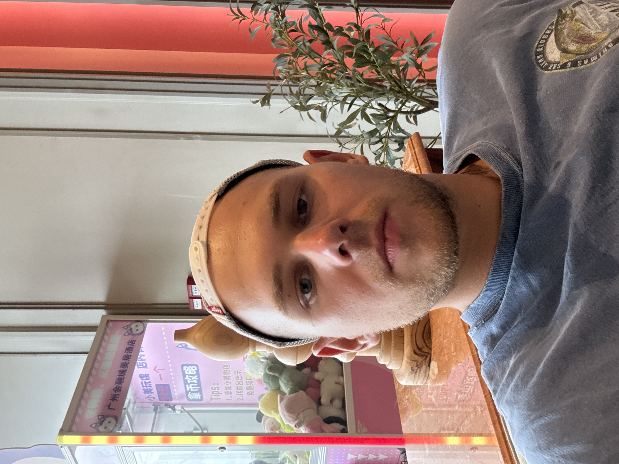 A young Caucasian man with light brown hair and a stubble beard is pictured in a close-up, selfie-style shot. He is wearing a light blue t-shirt with a circular emblem on the right side of his chest, depicting a fish and text that reads "JUAN RIVERS" and "SANTA ROSA". He has blue eyes and is wearing a light-colored brimmed hat. The background features a claw machine filled with stuffed animals, a wooden decorative item, and red and orange vertical blinds. The claw machine has Chinese text on it. The man is seated at a table with a marble-like surface, and to the right, a potted plant with green leaves is visible. The scene appears to be indoors, possibly in a hotel lobby or entertainment area in Huangpu District, China.