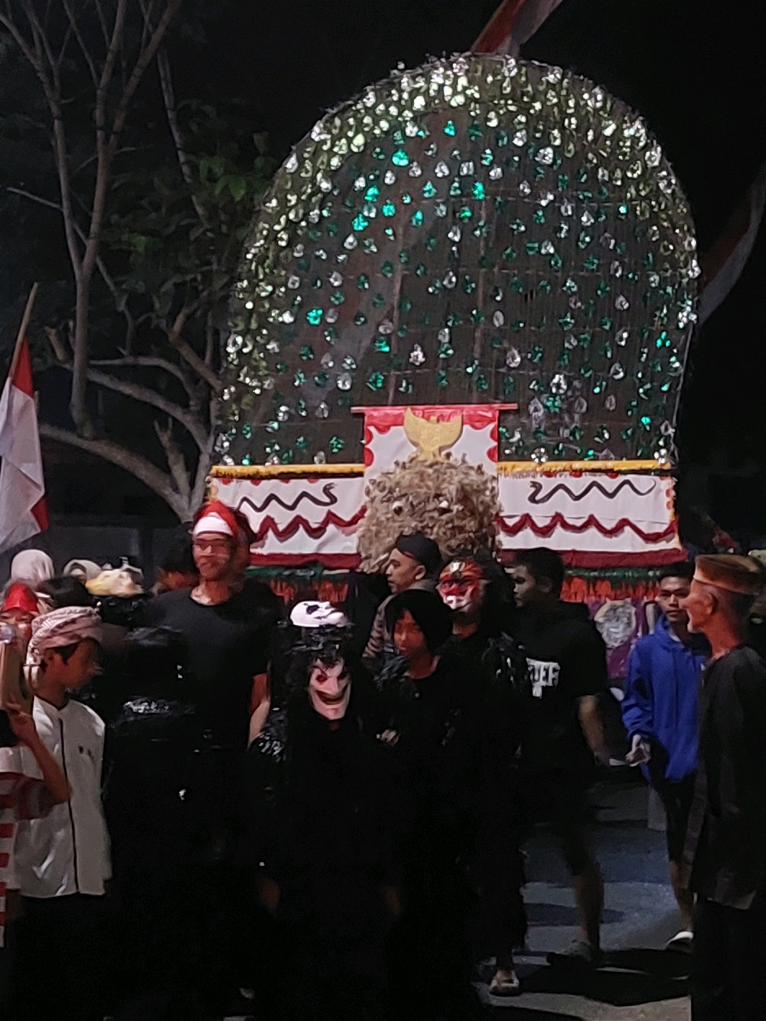 In the city of Ajung, Indonesia, a nighttime outdoor gathering is depicted. The central focus is a large, elaborate structure adorned with numerous small, reflective elements, some appearing green, attached to a grid-like surface. This structure is elevated and appears to be part of a procession or display. Below it, a decorated platform features a Garuda emblem, a symbol of Indonesia, and a wavy, serpent-like motif.
A group of people are visible, some wearing masks or traditional attire, suggesting a cultural event or celebration. One individual in the foreground wears a red Santa hat and glasses, facing towards the viewer. Several others are dressed in dark clothing, with some wearing masks, including one with a white face and striking features. Another person in the foreground wears a white and red striped shirt. The environment is dark, with the lighting primarily emanating from the illuminated structure and possibly portable lights. Trees with visible branches are present in the background, along with a partial view of a red and white flag, likely the Indonesian flag. The ground appears to be paved.