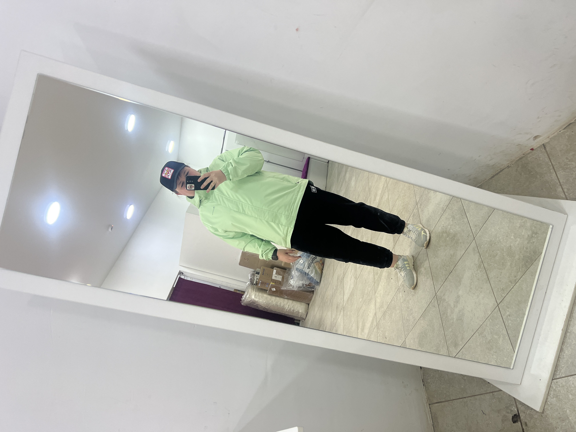 A person is standing in front of a full-length mirror, taking a selfie with a mobile phone. The person is wearing a black baseball cap, a light green jacket, black pants, and light-colored sneakers. The mirror is framed in white and is placed against a white wall. The reflection shows a room with boxes and rolled-up rugs. The floor is tiled with light grey tiles. A partially visible purple curtain can be seen in the background. The photo was taken in Chaikovsky, Russia.