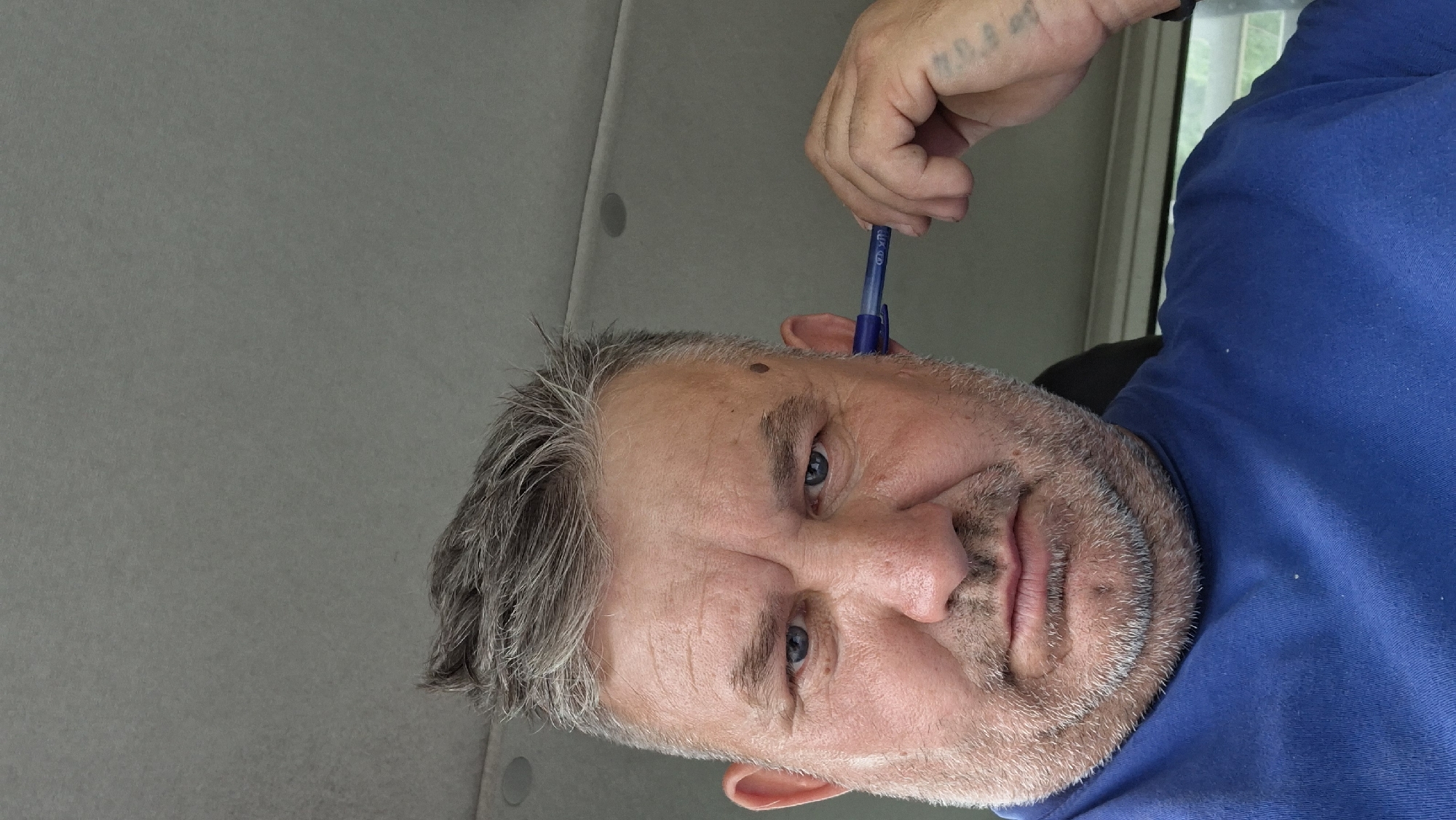 A middle-aged man with graying hair and a beard is pictured from the chest up, looking directly at the camera. He is wearing a blue t-shirt. A blue pen is held to his right ear. The background appears to be the interior of a vehicle, with the ceiling visible and a hint of greenery through a window. There is no identifiable information or context that would place the image specifically in Hîncești, Moldova.