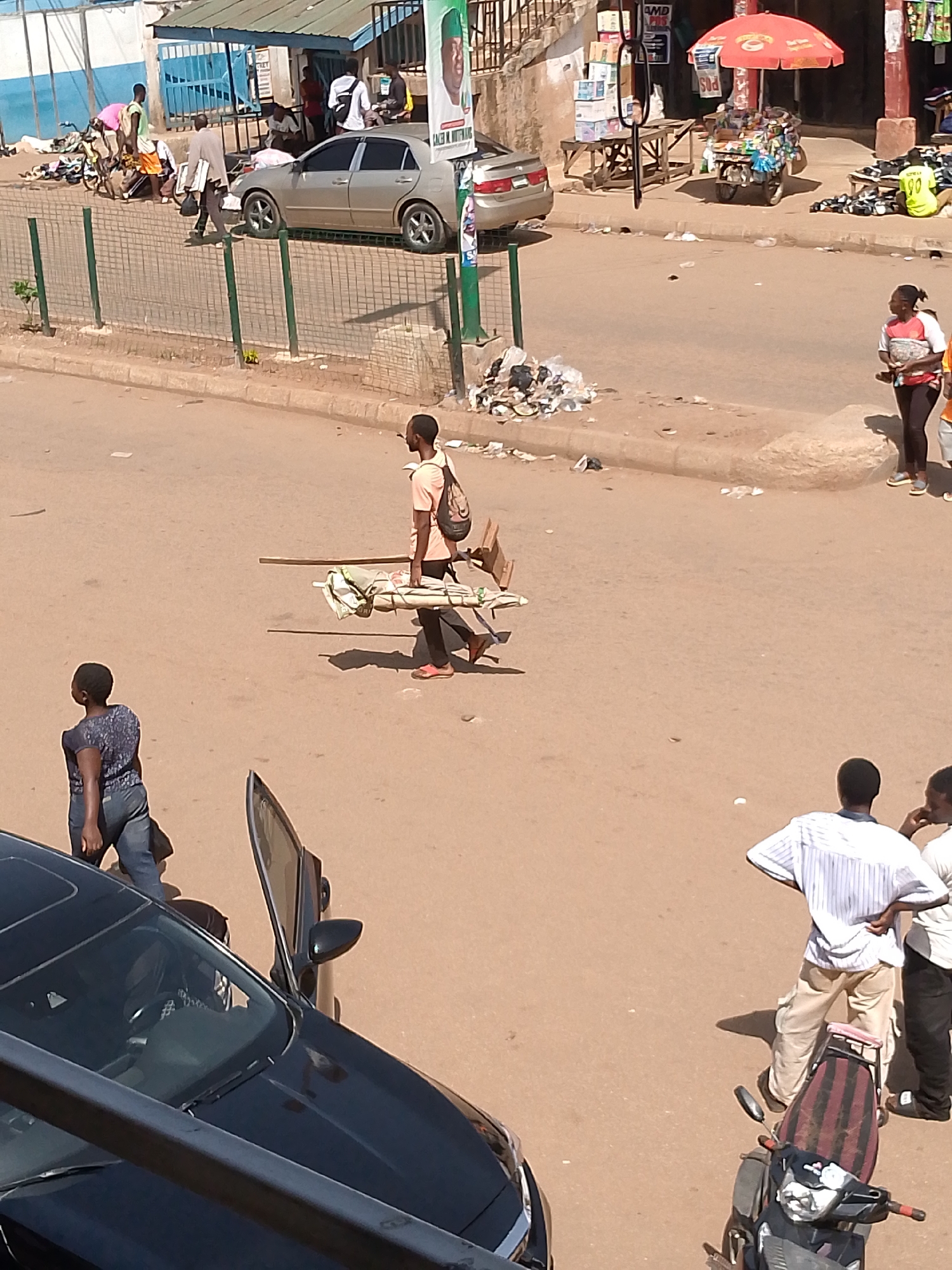 The image depicts a street scene in Bukuru, Nigeria. The setting is a dusty, sunlit street with a mix of pedestrian and vehicular traffic. In the foreground, a black car with an open door is parked, partially obstructing the view. A woman in jeans and a dark top is walking towards the car. In the middle ground, a man is carrying a load balanced on his shoulders, appearing to transport items across the street. Two men are conversing near a parked motorcycle in the lower right of the image. In the background, there are shops and market stalls, with various people milling about. A silver car is parked along the side of the road. Rubbish is scattered along the side of the road. The overall impression is of a busy, typical urban street scene in Nigeria.