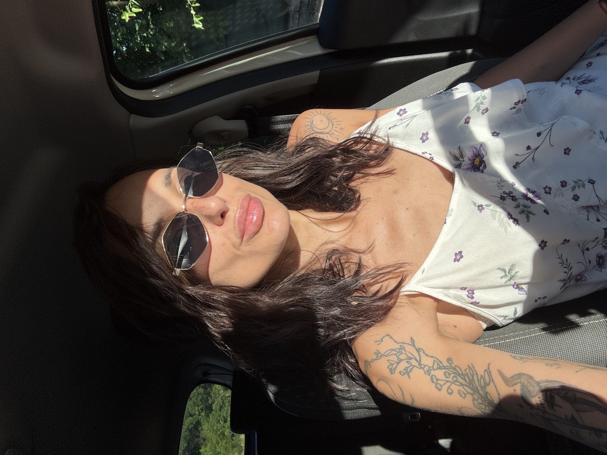 A woman is sitting in the driver's seat of a car. She is wearing sunglasses and a white dress with a floral pattern. Her hair is dark brown and wavy. She has tattoos on her left arm and shoulder. The car is parked outdoors, with trees visible through the windows. Sunlight is streaming into the car, creating shadows on her face and body. The location is the city of Pleven, Bulgaria.