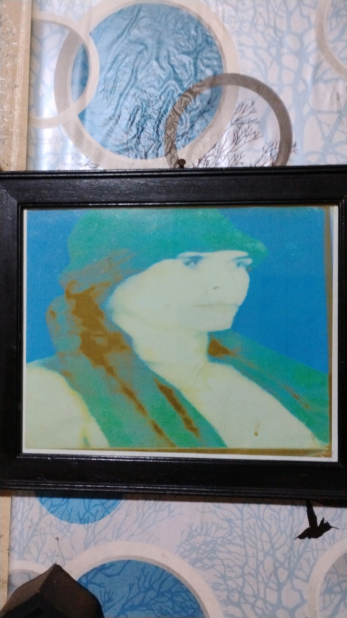 The image shows a framed portrait of a person wearing a hat and scarf against a blue background. The portrait is rendered in abstract, impressionistic style with broad strokes of color. The frame is black and appears to be made of wood. The portrait is hanging on a wall with a patterned wallpaper. The wallpaper has a blue and white design with abstract circular shapes and branch-like patterns. A small silhouette of a bird in flight is visible in the lower right corner of the image, on the wallpaper. The image is taken from a slightly high angle, and part of a dark wooden object is visible in the lower left corner. The overall setting appears to be indoors, possibly a home or a small establishment. There is no specific identifiable landmark or clue that places this image in West Jakarta, Indonesia.