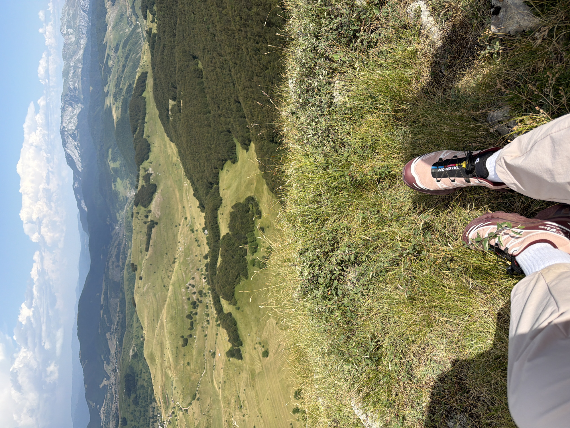 The image captures a person's view from a grassy hilltop overlooking a vast, undulating landscape. In the foreground, the person's legs and feet are visible, clad in light-colored pants and athletic shoes, one of which is a Salomon brand trail runner with a distinctive lacing system. The shoes are adorned with a few sprigs of green foliage. The ground beneath is covered in dry, yellow-tinged grass with scattered small green plants and some rocky outcrops.
The midground features rolling green hills, interspersed with patches of dark green trees, likely a forest, and some lighter green fields that appear to be meadows or agricultural land. A winding road is faintly visible in one of the fields. Further in the distance, layers of mountains recede into the horizon, their peaks varying in texture and color, with some showing rocky, lighter-colored formations against a backdrop of hazy blue mountain ranges. The sky is a bright blue with scattered white, fluffy clouds.
This scene is consistent with a location within Trnovo Municipality (FBiH), Bosnia and Herzegovina, known for its mountainous terrain and natural beauty, particularly areas suitable for hiking and outdoor activities such as the Bjelašnica mountain region, which is part of this municipality and offers similar panoramic views.