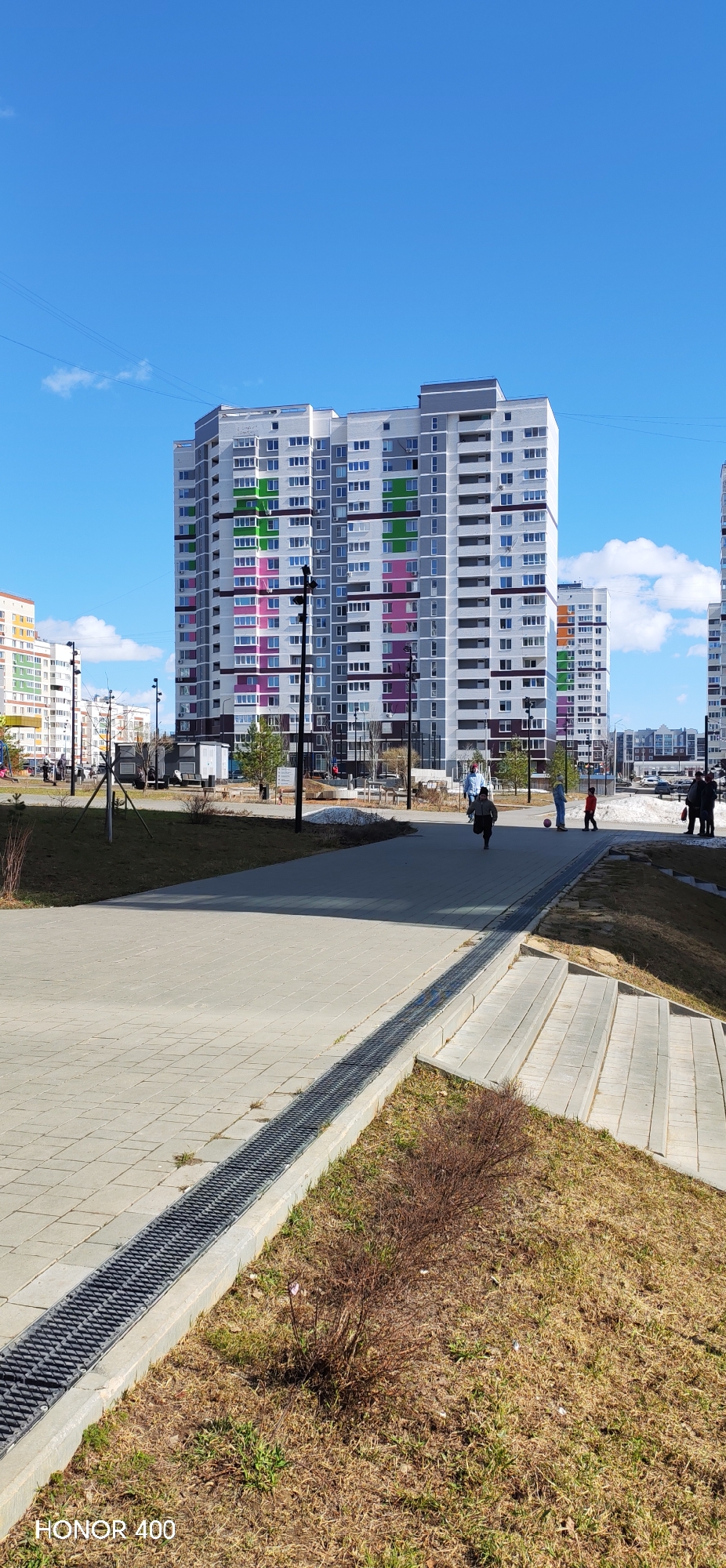 The image depicts a paved walkway in a modern urban area, likely within the city of Izhevsk, Russia, given the contemporary architecture and general environment.
The walkway is constructed from square, light-colored paving stones and features a drainage grate running along one edge. Adjacent to the walkway are grassy areas with patches of dry vegetation, suggesting early spring or late fall. Steps lead down from the walkway to a lower level.
In the background are several tall, multi-story residential buildings characterized by a modern design with rows of windows and colorful accent panels.
Several people are present. A child is running on the walkway, while other individuals stand in small groups or walk leisurely. Some people seem to be socializing near a grassy area.
The sky is blue with scattered white clouds, indicating a clear day. Street lamps are visible along the walkway.